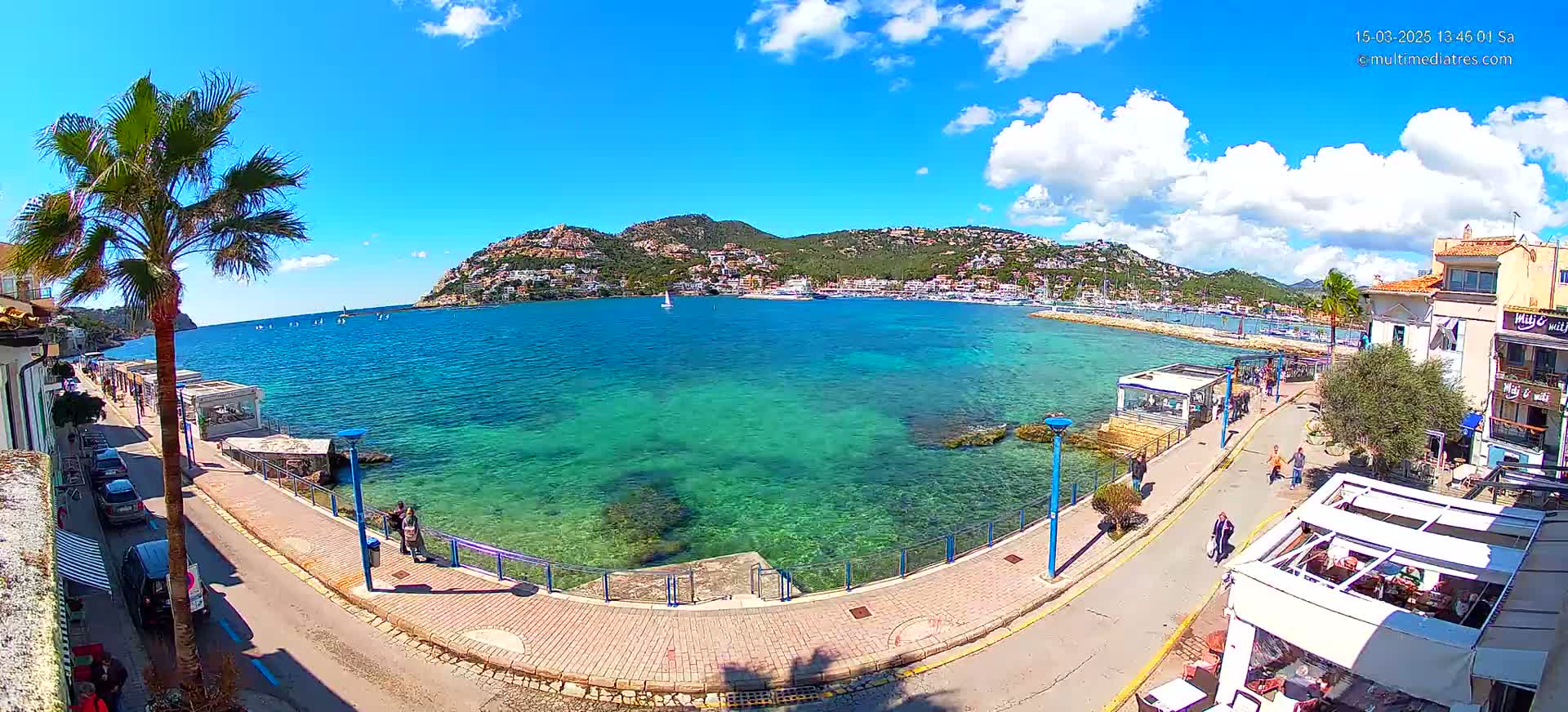 A sunny day with partly cloudy skies shows a clear, turquoise harbor with boats and buildings lining a paved waterfront path.