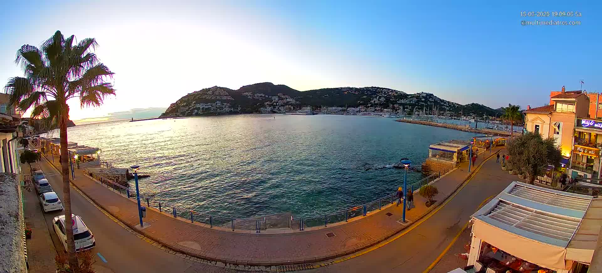 A calm, sun-lit harbor scene shows a waterfront promenade with parked cars, restaurants, and people walking, overlooking a bay with boats and a hill in the background.