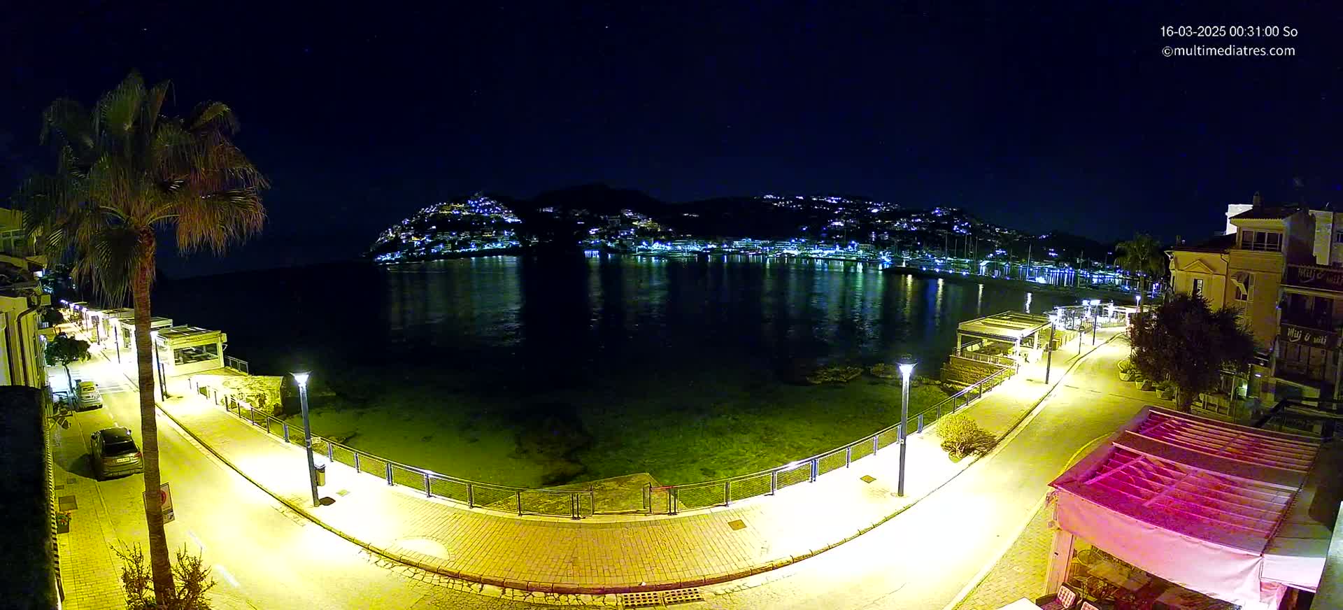 A nighttime, waterfront scene shows a paved walkway illuminated by streetlights, curving around a calm bay with a hilltop town sparkling with lights across the water.
