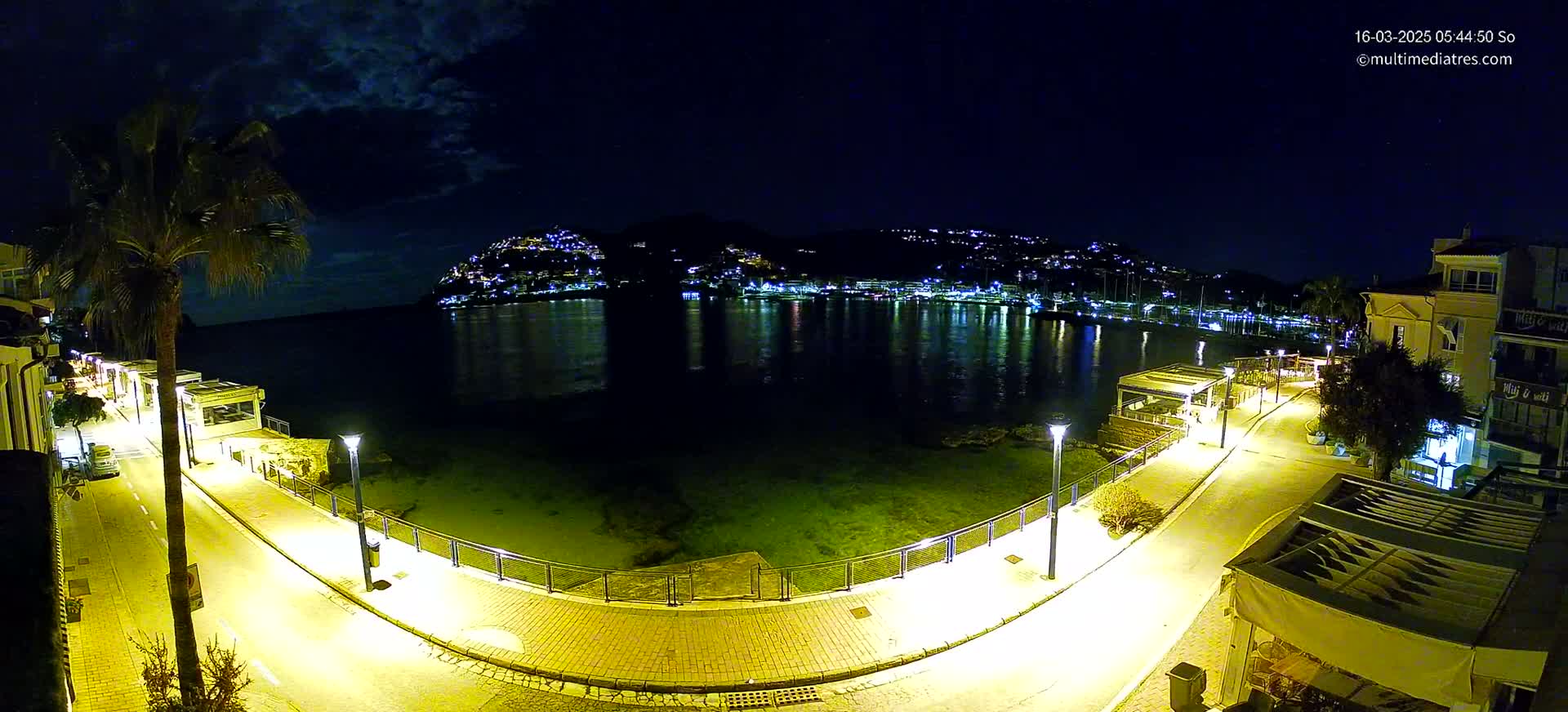 A nighttime view of a waterfront promenade illuminated by streetlights, overlooking a calm bay with a hilltop town reflecting in the water under a partly cloudy sky.