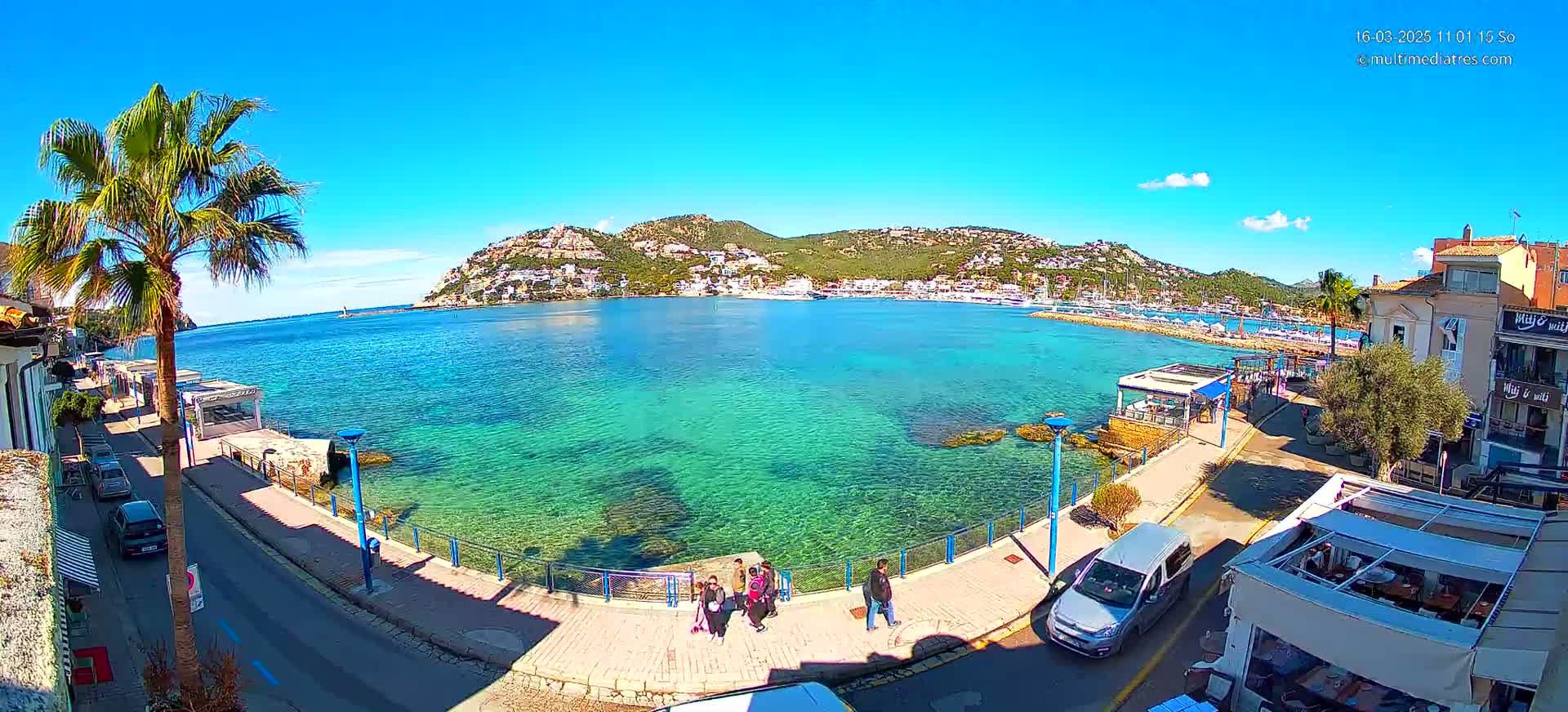 A sunny day reveals a clear, turquoise bay with a town nestled against a hillside, cars parked along a seaside promenade, and several people walking nearby.