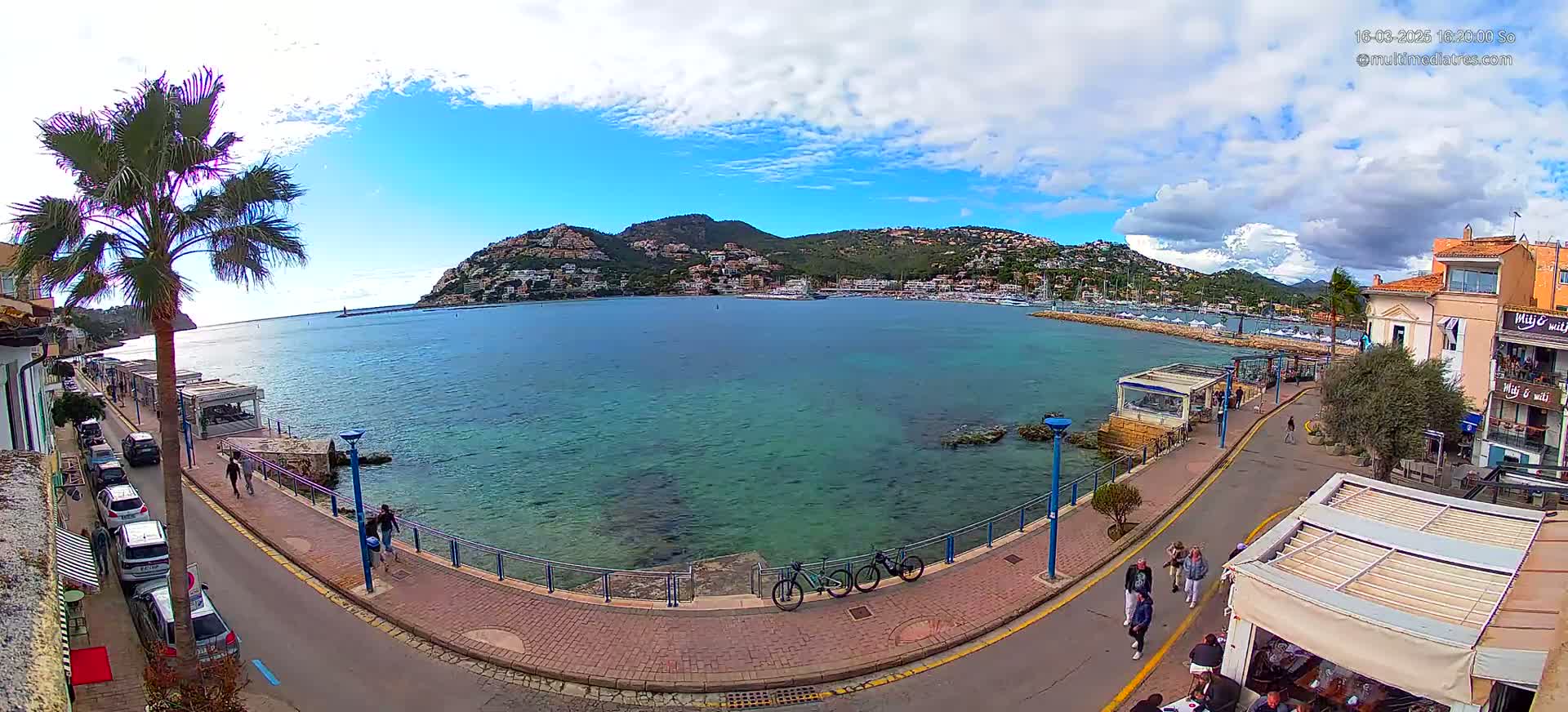 A partly cloudy day shows a waterfront town with a calm, turquoise bay, a hillside town across the water, and people walking along a paved path.