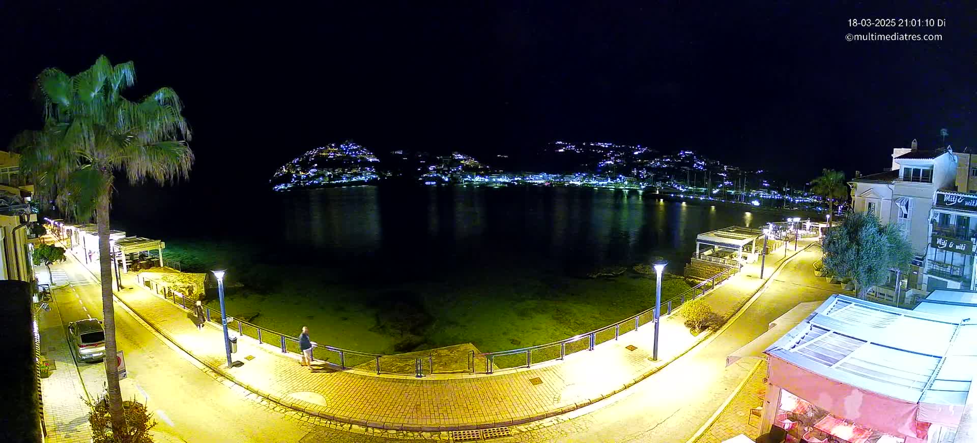 A nighttime, seaside view shows a paved walkway lit with yellow lights curving around a calm bay, with buildings and a hill dotted with lights in the background.