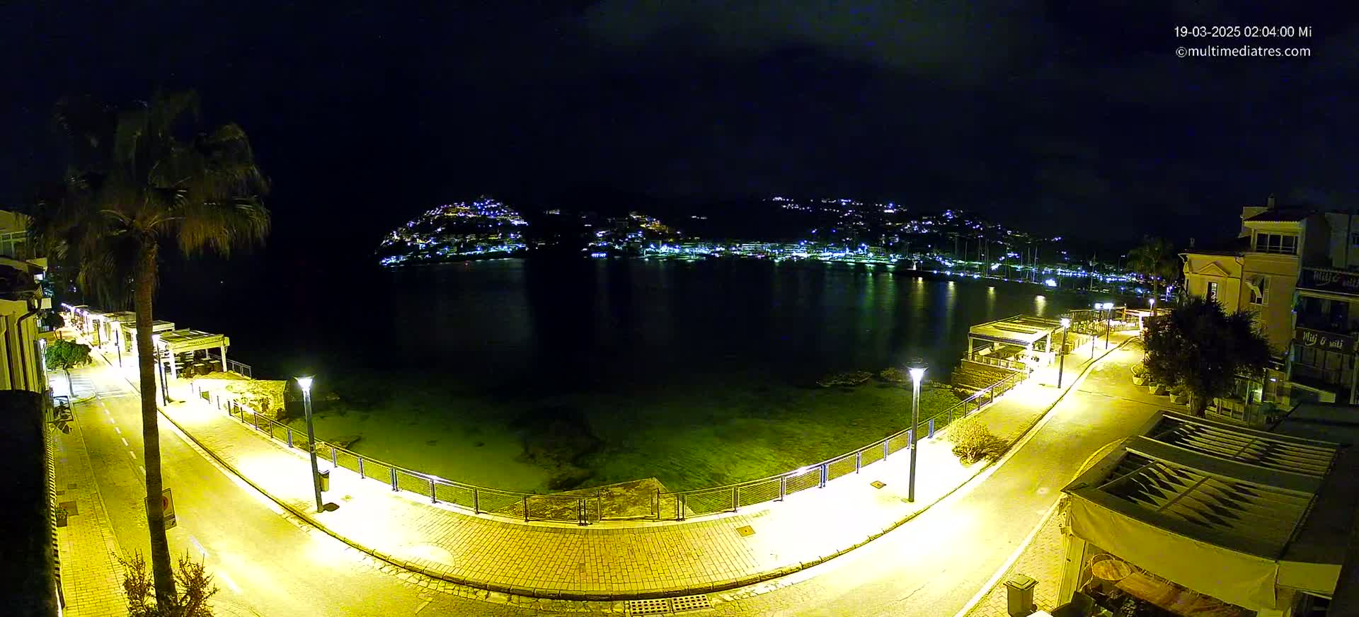 A nighttime, illuminated waterfront scene shows a calm bay with a hilltop town across the water, and a paved walkway curving along the shoreline.