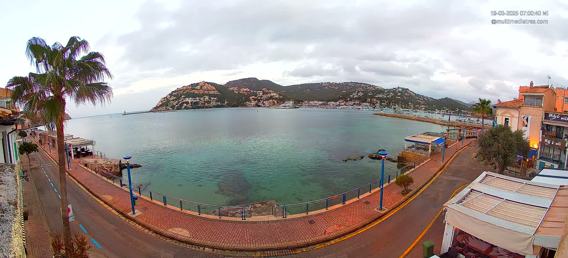 A calm bay with clear, shallow water is seen from a waterfront promenade on an overcast day, with a hill covered in buildings in the background.