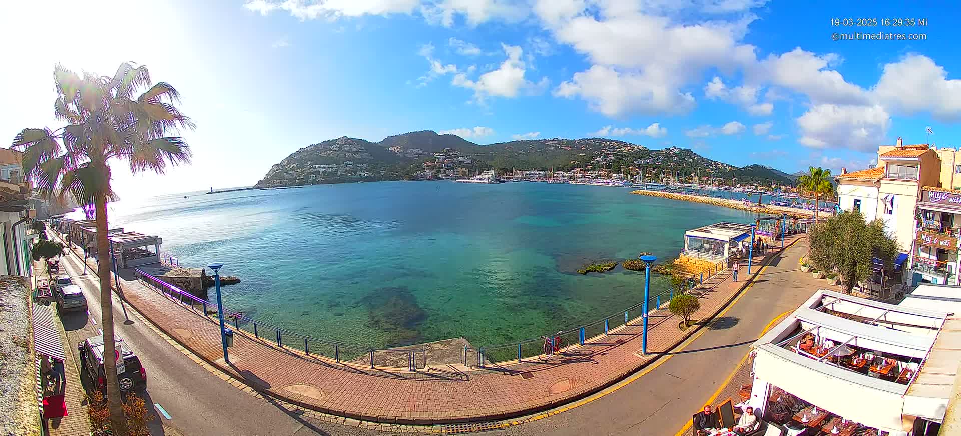 A sunny day reveals a seaside town with clear turquoise water, a promenade lined with restaurants, and a hill covered in buildings in the background.