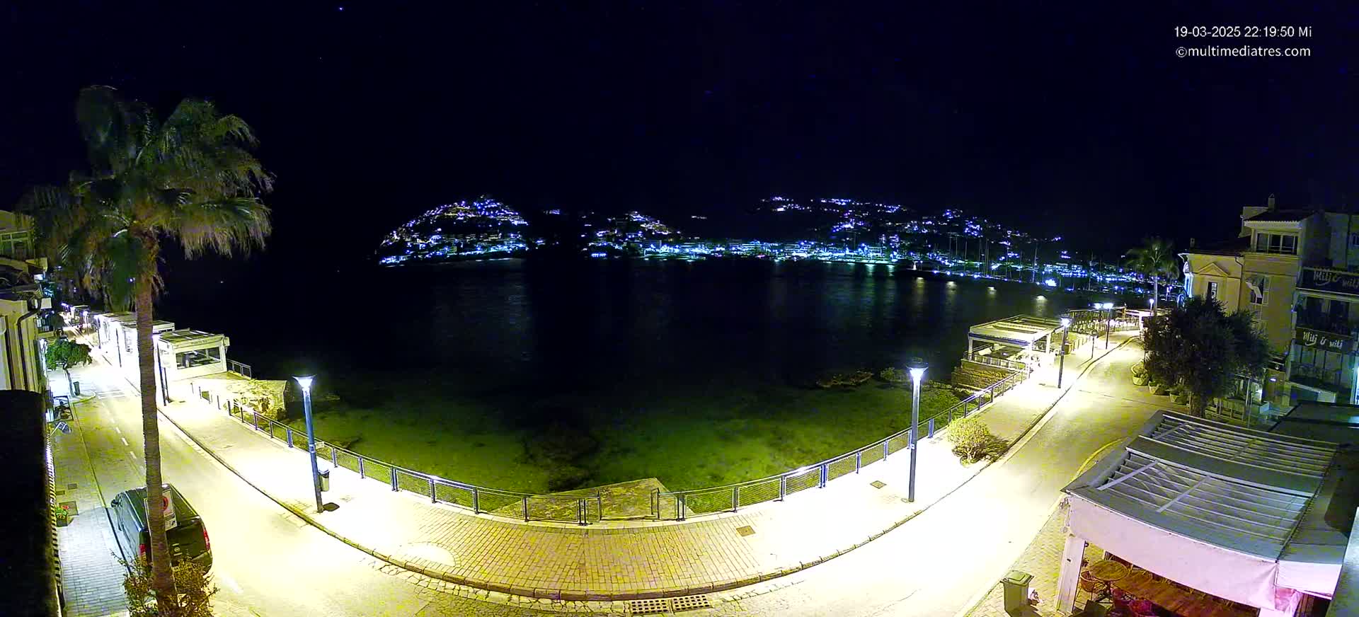 A nighttime, coastal scene shows a calm body of water reflecting the lights of a town built on a hillside, with a paved walkway curving along the shore illuminated by streetlights.