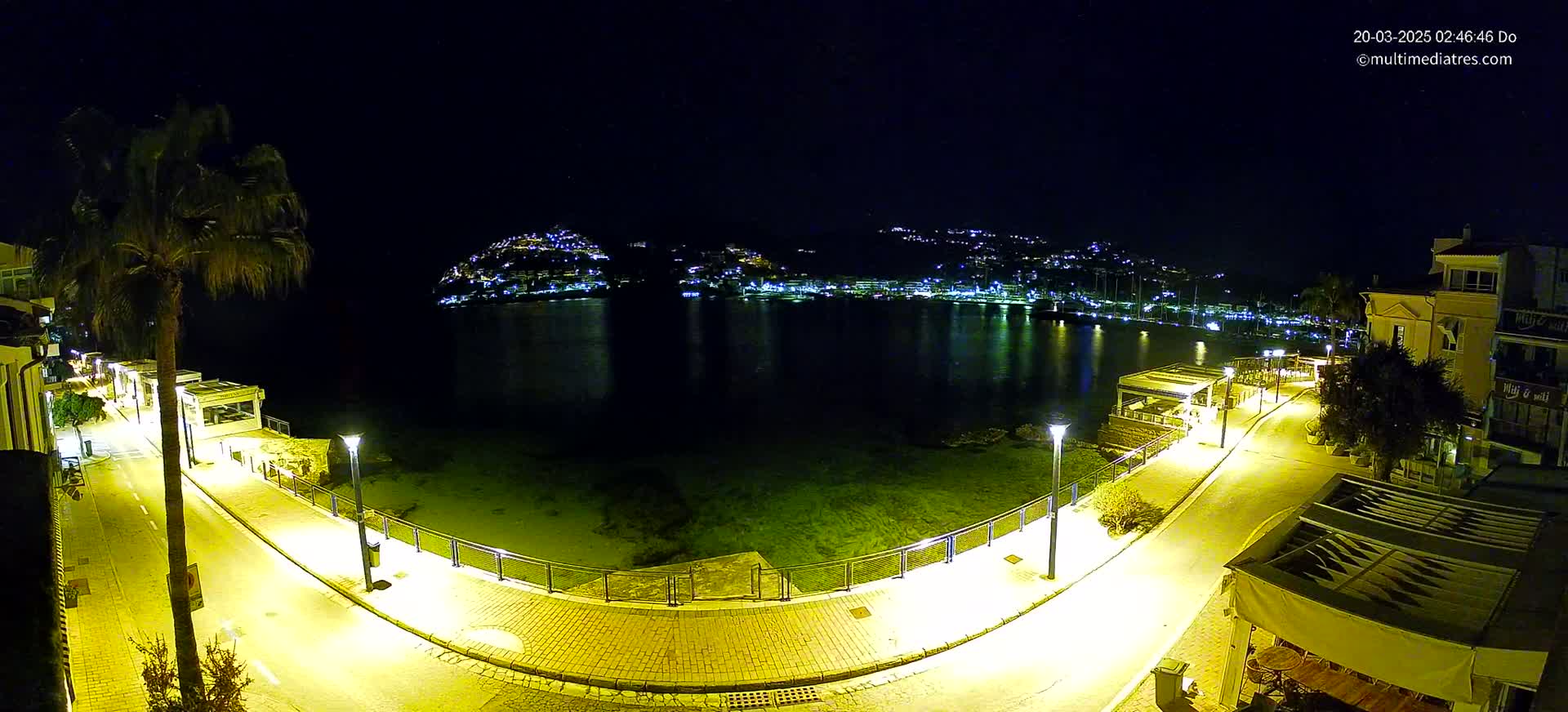 A nighttime, aerial view shows a calm body of water reflecting the lights of a town nestled on a hillside, with a brightly lit paved walkway curving along the waterfront.