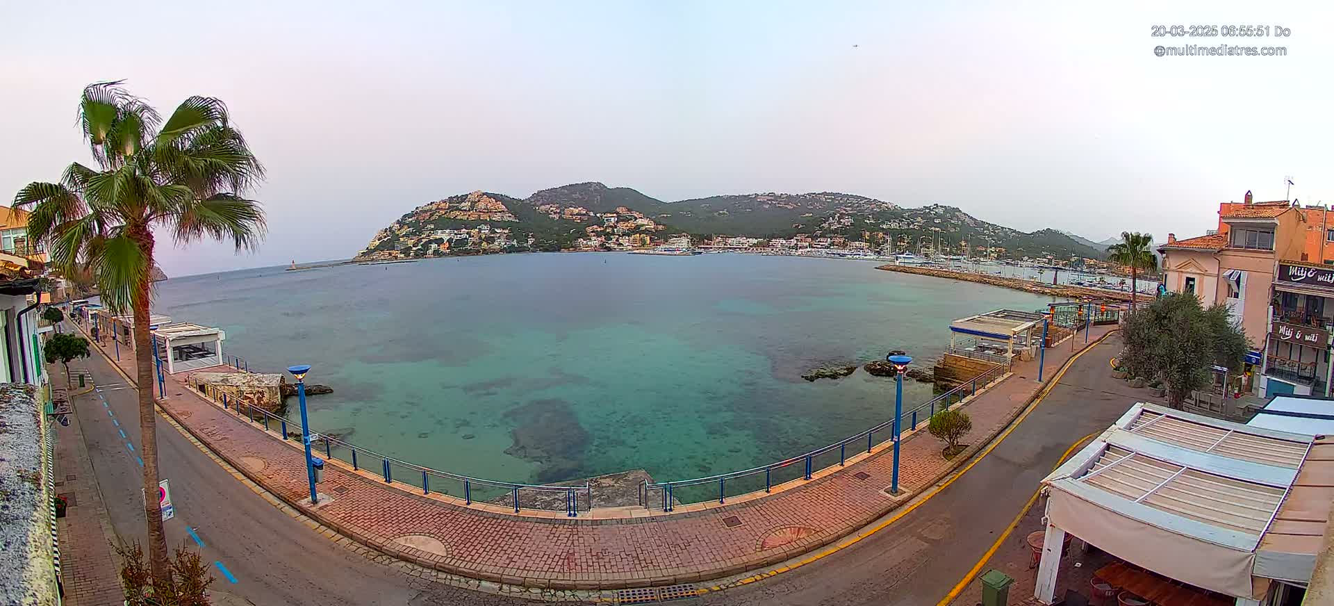 A paved waterfront path curves around a calm, clear turquoise bay with a hill covered in buildings in the background under an overcast sky.
