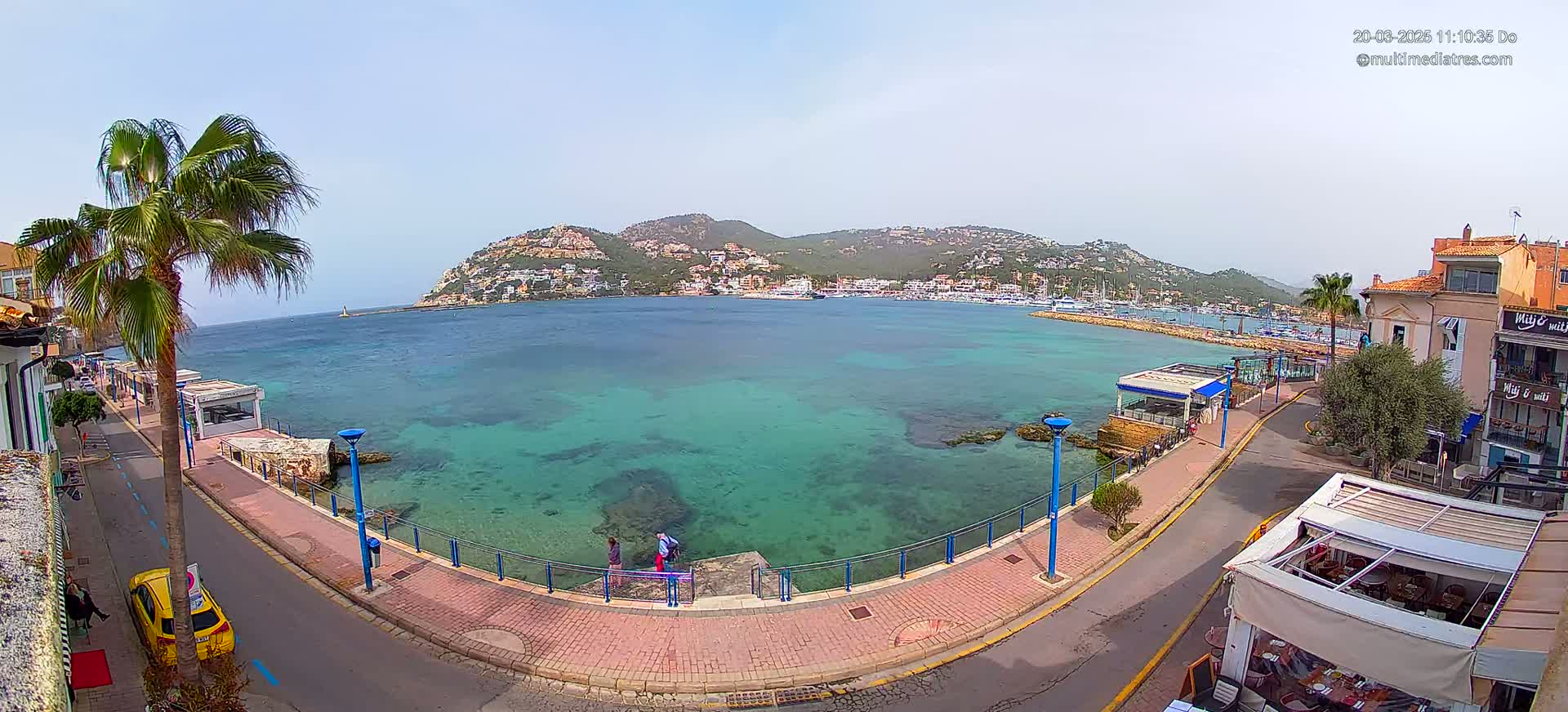 A clear, sunny day reveals a coastal town's waterfront, showcasing a calm, turquoise bay surrounded by buildings and a hill covered in houses.