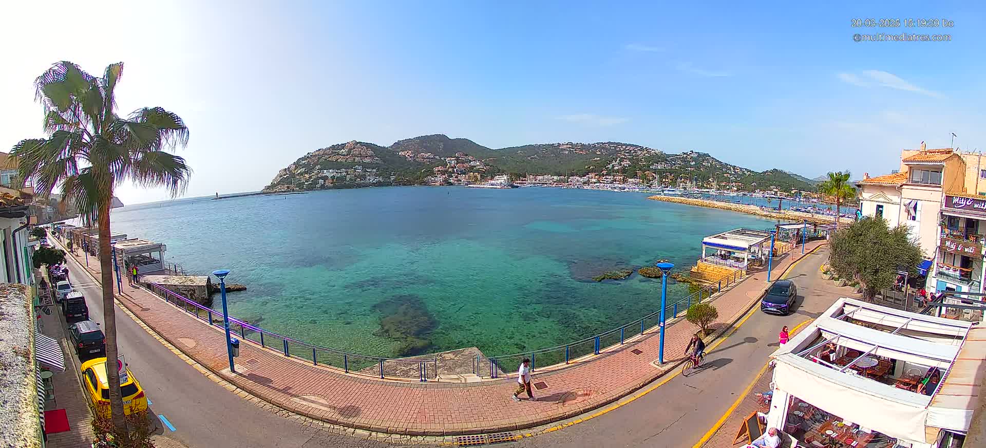A clear sunny day overlooks a calm, shallow, teal-colored bay with a town and hillside buildings in the background,  a paved walkway, and several people walking and cycling along the waterfront road.