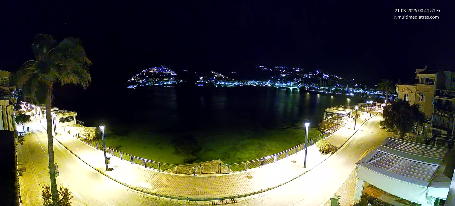 A nighttime, high-angle view shows a coastal town's illuminated waterfront promenade curving around a calm bay, with a hilltop town glowing with lights in the background.