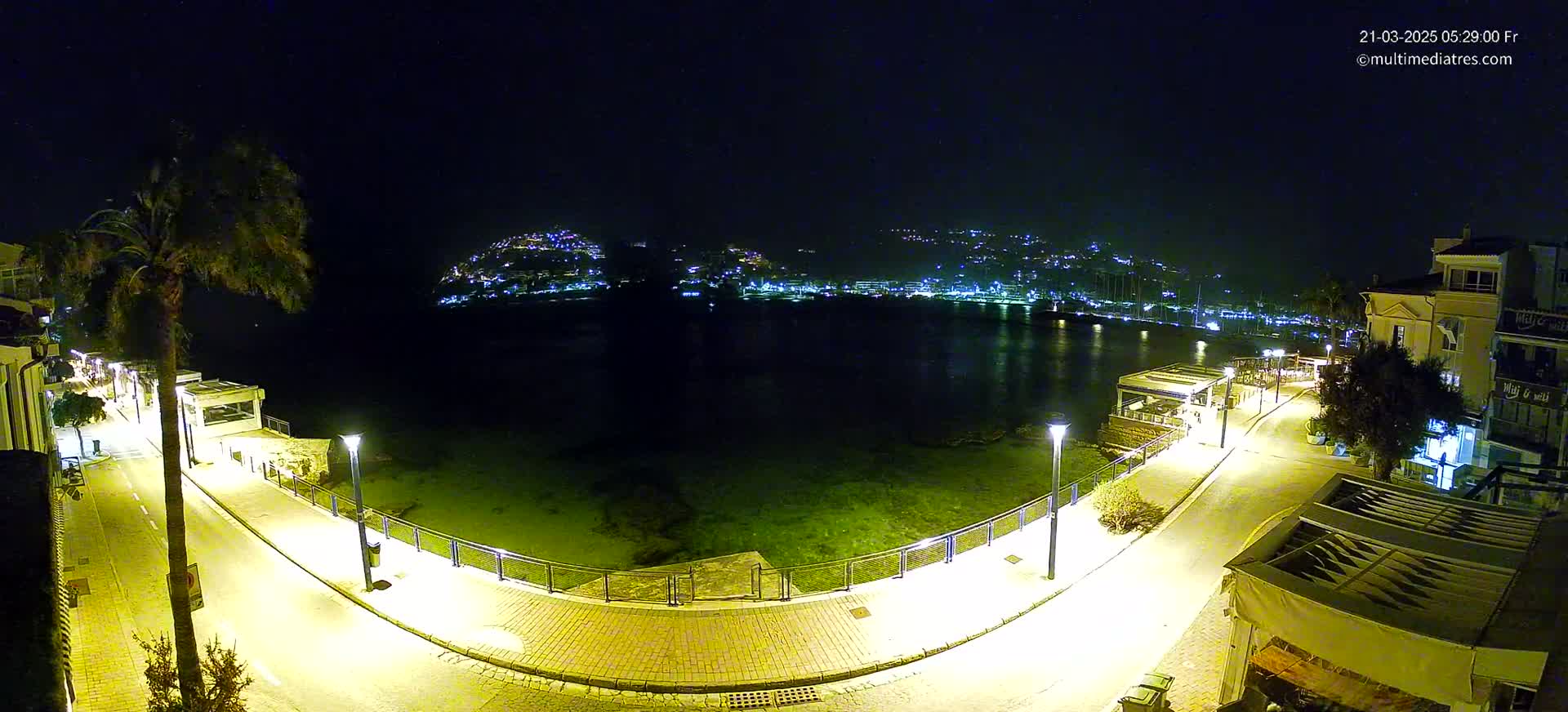 A nighttime, illuminated waterfront scene shows a calm body of water with a town's lights visible across it, a curved roadway with lamp posts and buildings lining it.