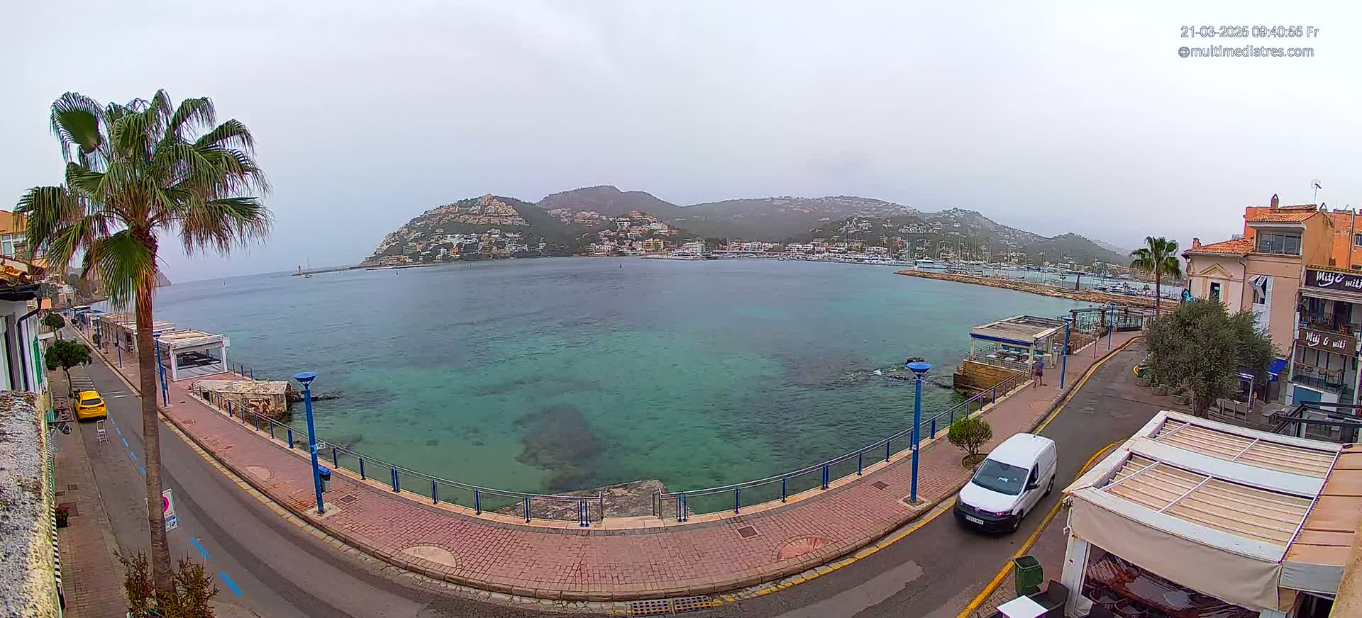 A hazy, overcast day reveals a calm bay with clear, turquoise water, bordered by a paved waterfront path and buildings, and hills in the background.