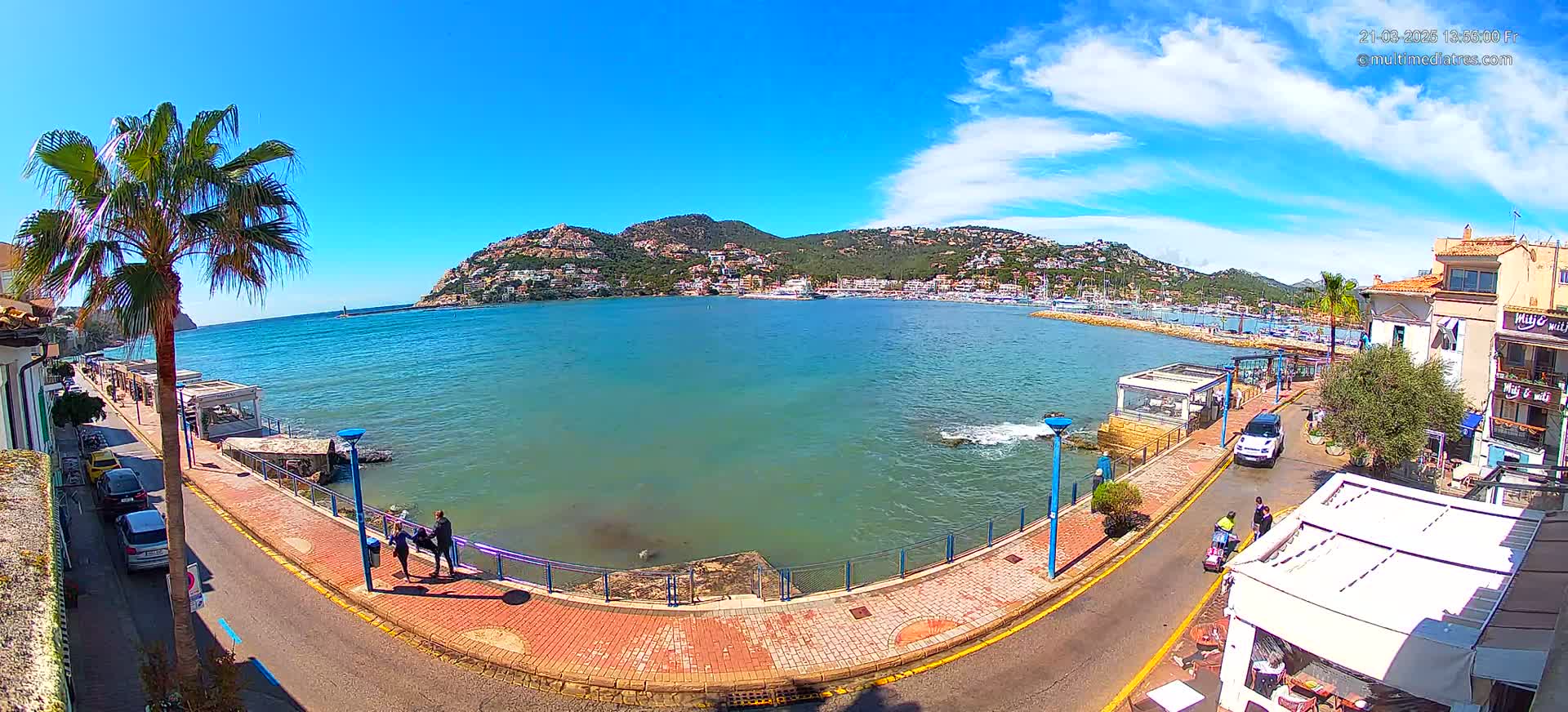 A seaside town with a calm bay, under a sunny sky with scattered clouds, is viewed from a high vantage point, showing a paved walkway, parked cars, and some people walking along the waterfront.