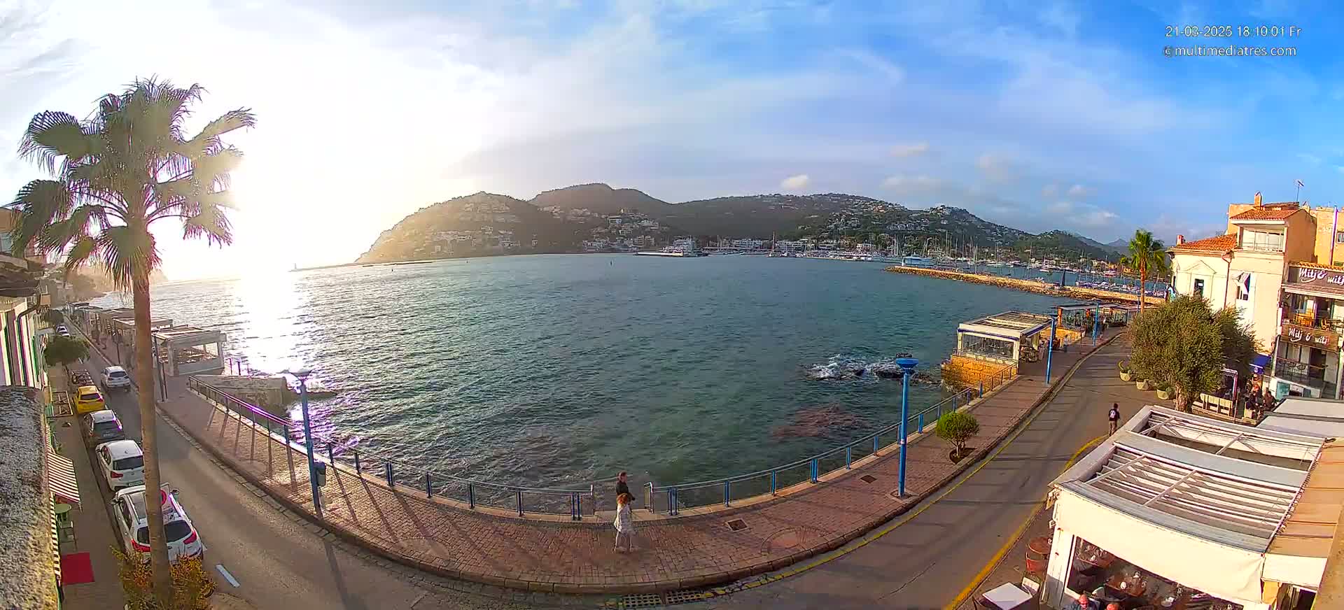 A seaside town at sunset, with a calm bay filled with boats and a paved walkway lined with buildings and palm trees.