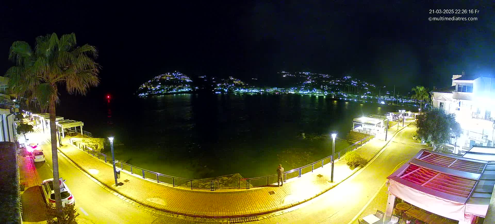 A nighttime view of a calm body of water with a town illuminated on the opposite shore, a paved walkway and several parked cars lining the water's edge.