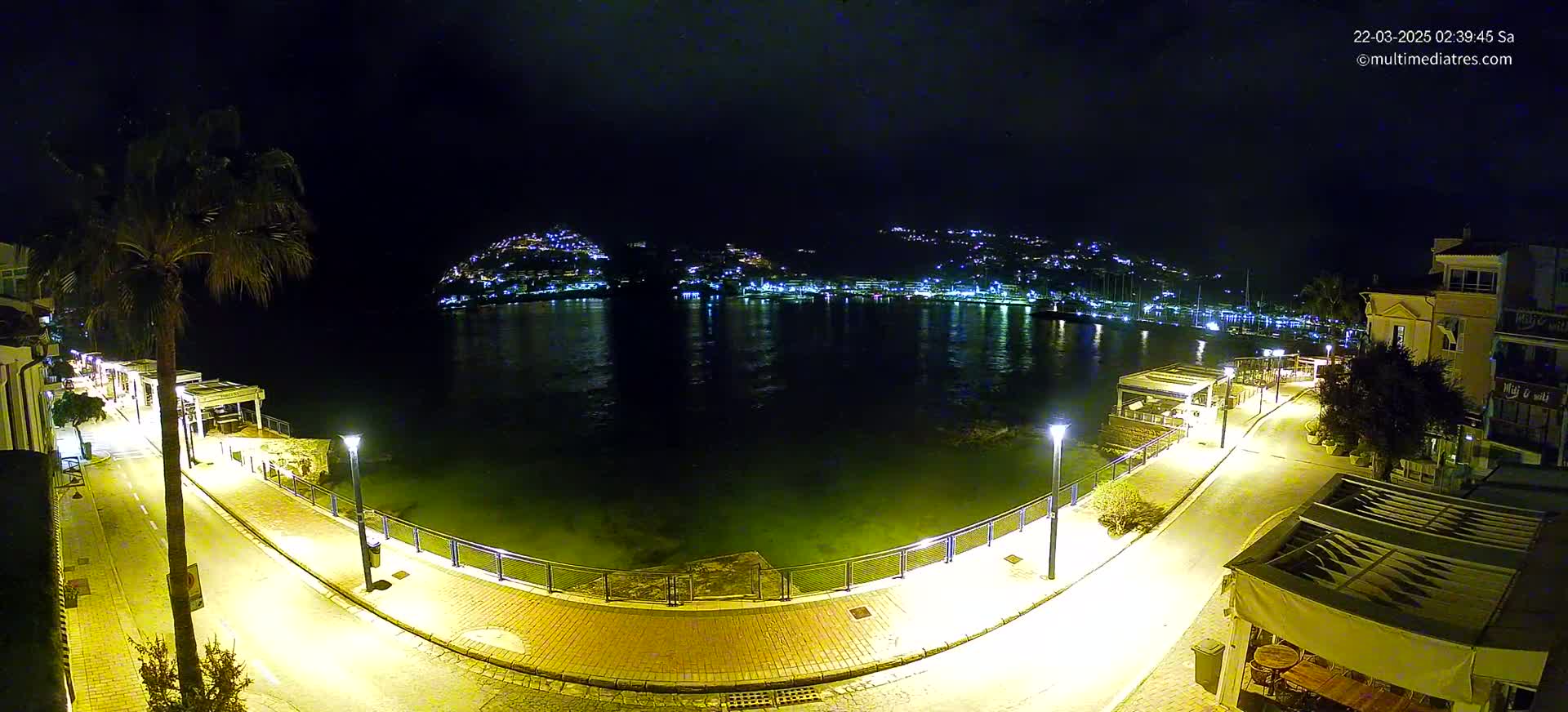 A nighttime view of a calm harbor, illuminated by streetlights, with a hilltop town glowing in the distance.