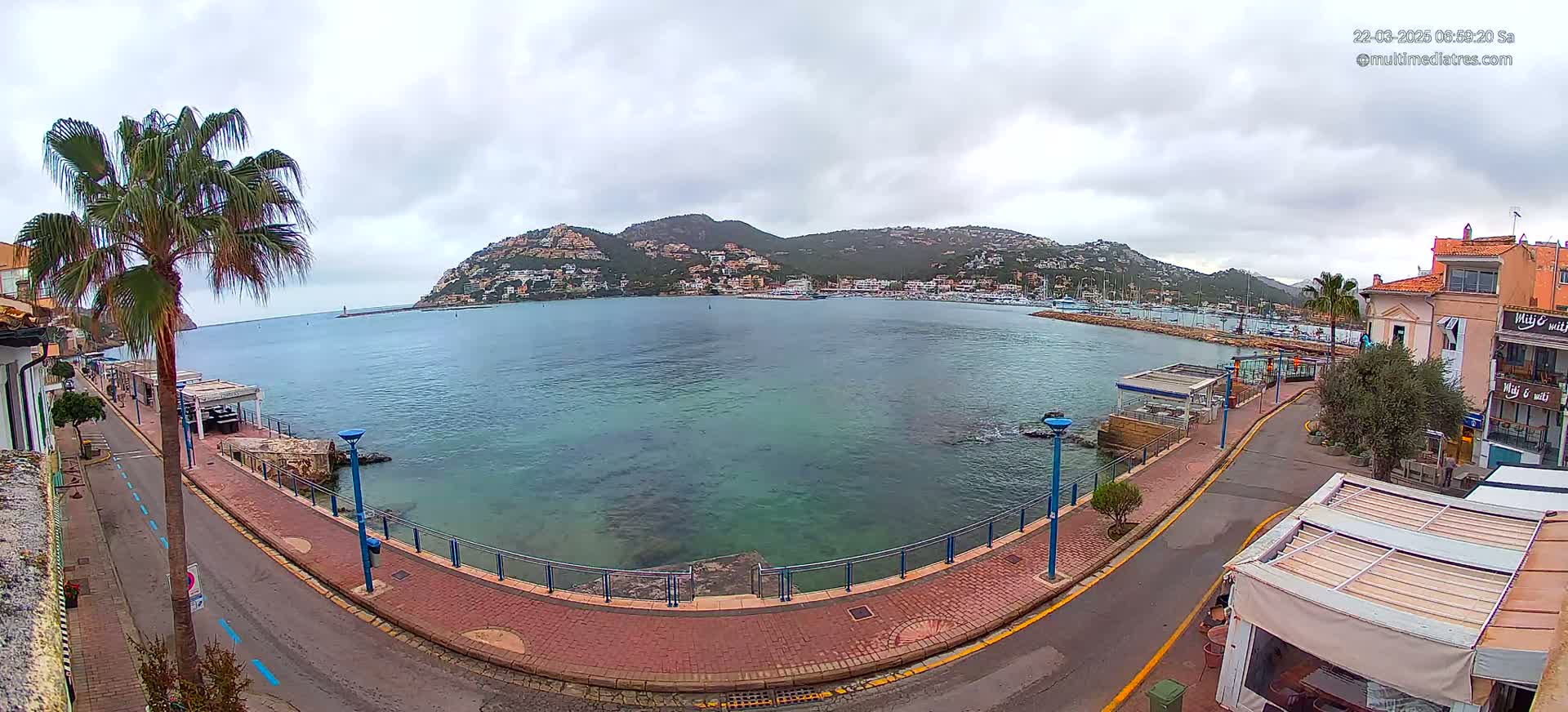 A calm, overcast day reveals a harbor town nestled at the base of a hill, with a paved walkway alongside the clear, teal water.