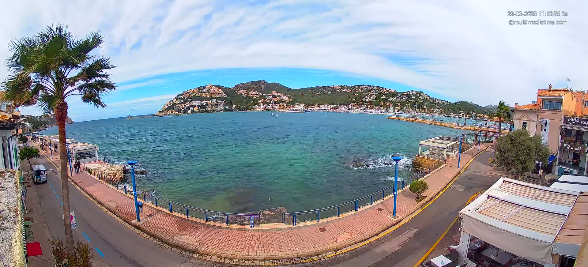A seaside town with palm trees lines a paved walkway overlooking a calm, blue bay and a hill covered with buildings under a partly cloudy sky.