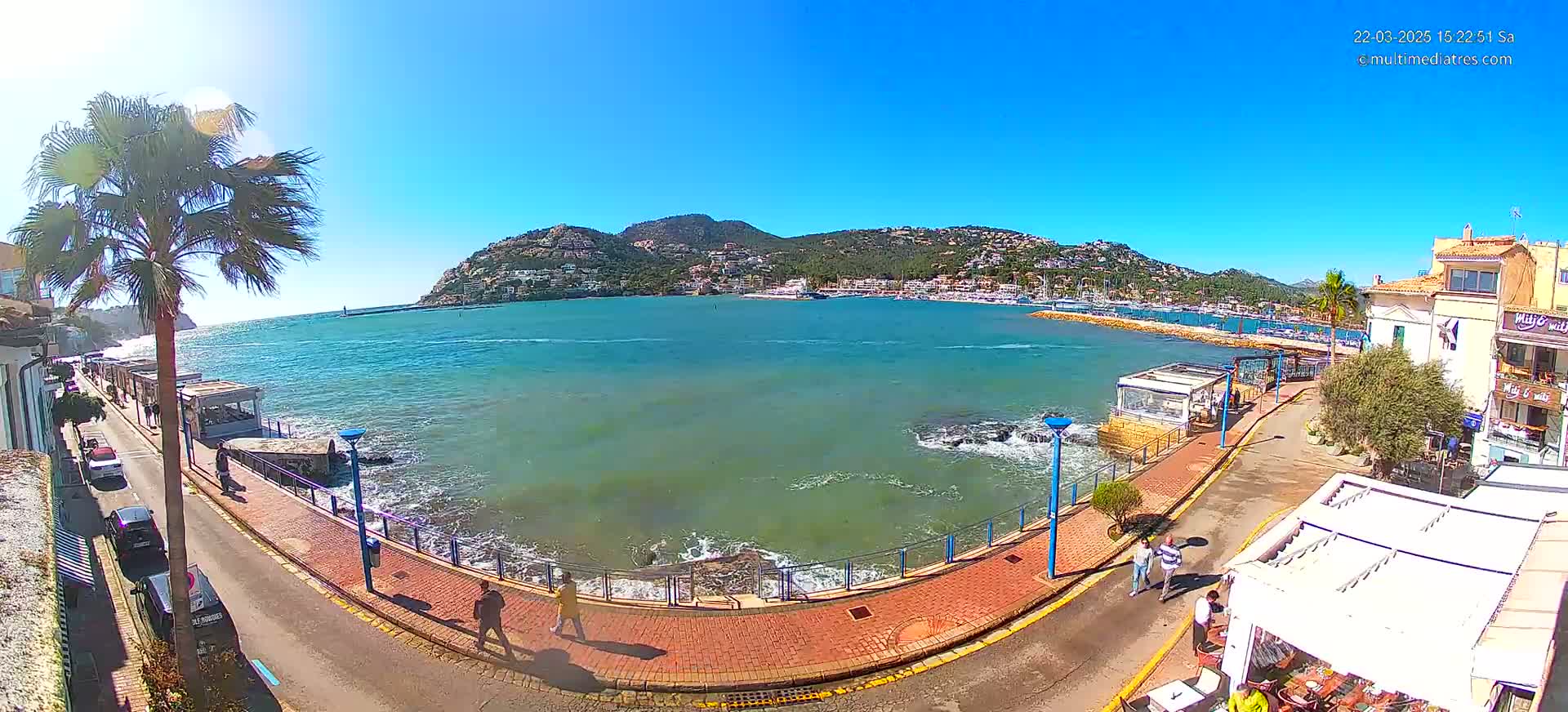 A sunny day reveals a coastal town with a harbor filled with boats, a brick walkway along the water's edge, and small waves crashing against rocks.