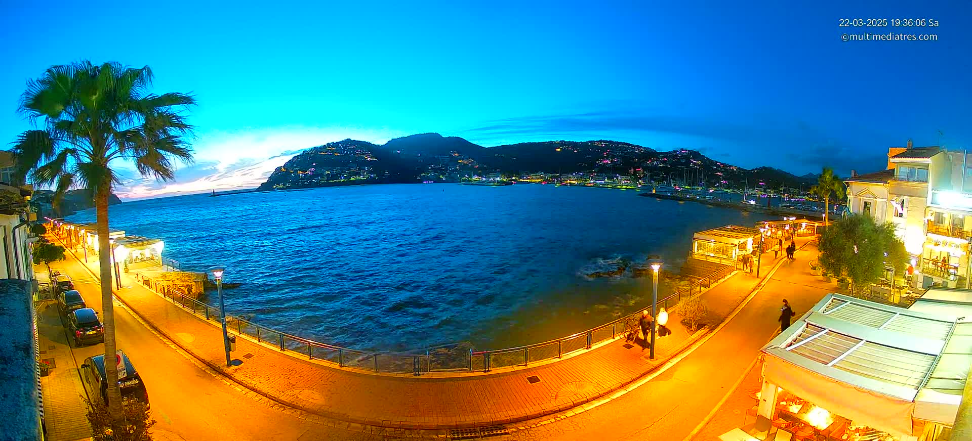 A nighttime seaside town scene shows a calm, dark blue body of water lapping against a paved walkway lined with lit buildings and palm trees, under a twilight sky.
