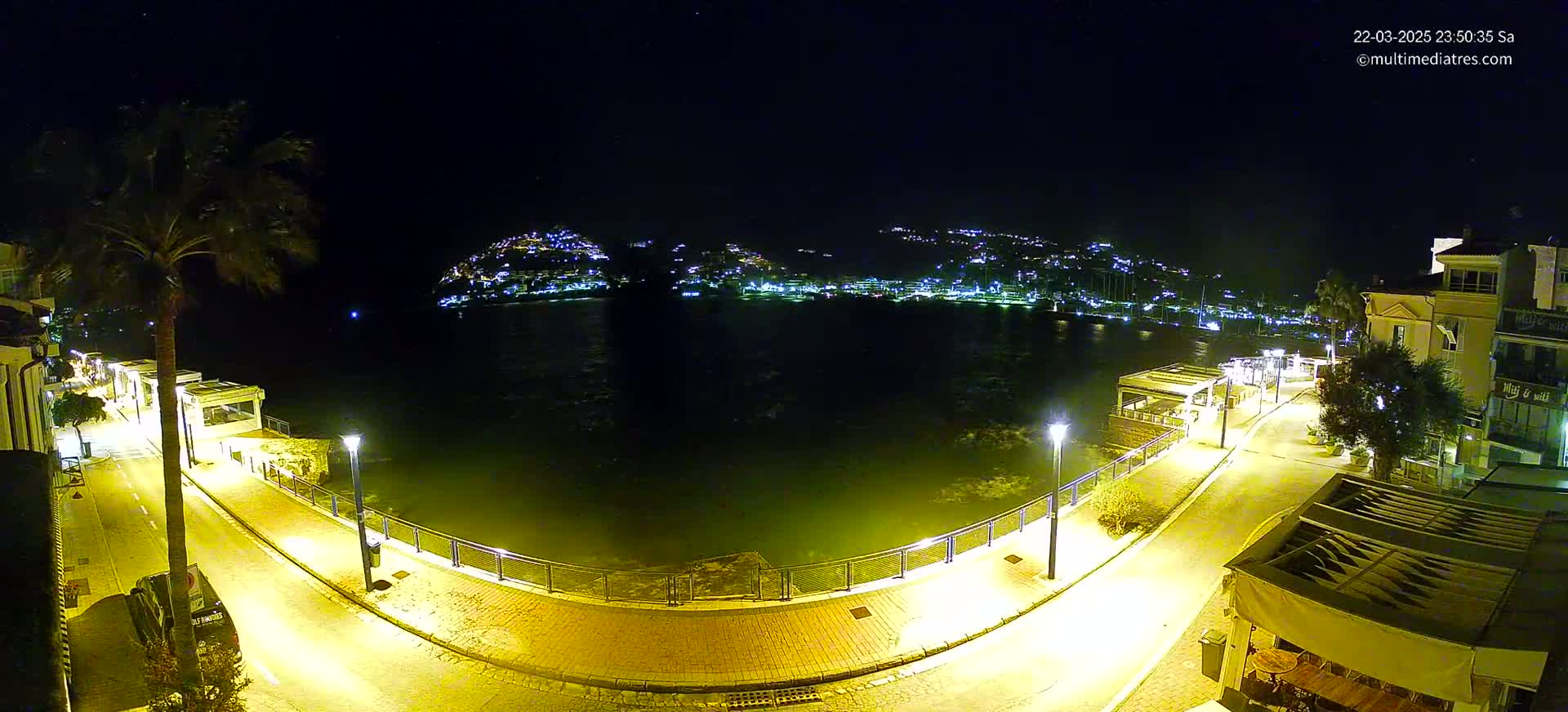 A nighttime view of a calm body of water, bordered by a paved walkway with streetlights illuminating buildings and vegetation, across from a hill dotted with lights.
