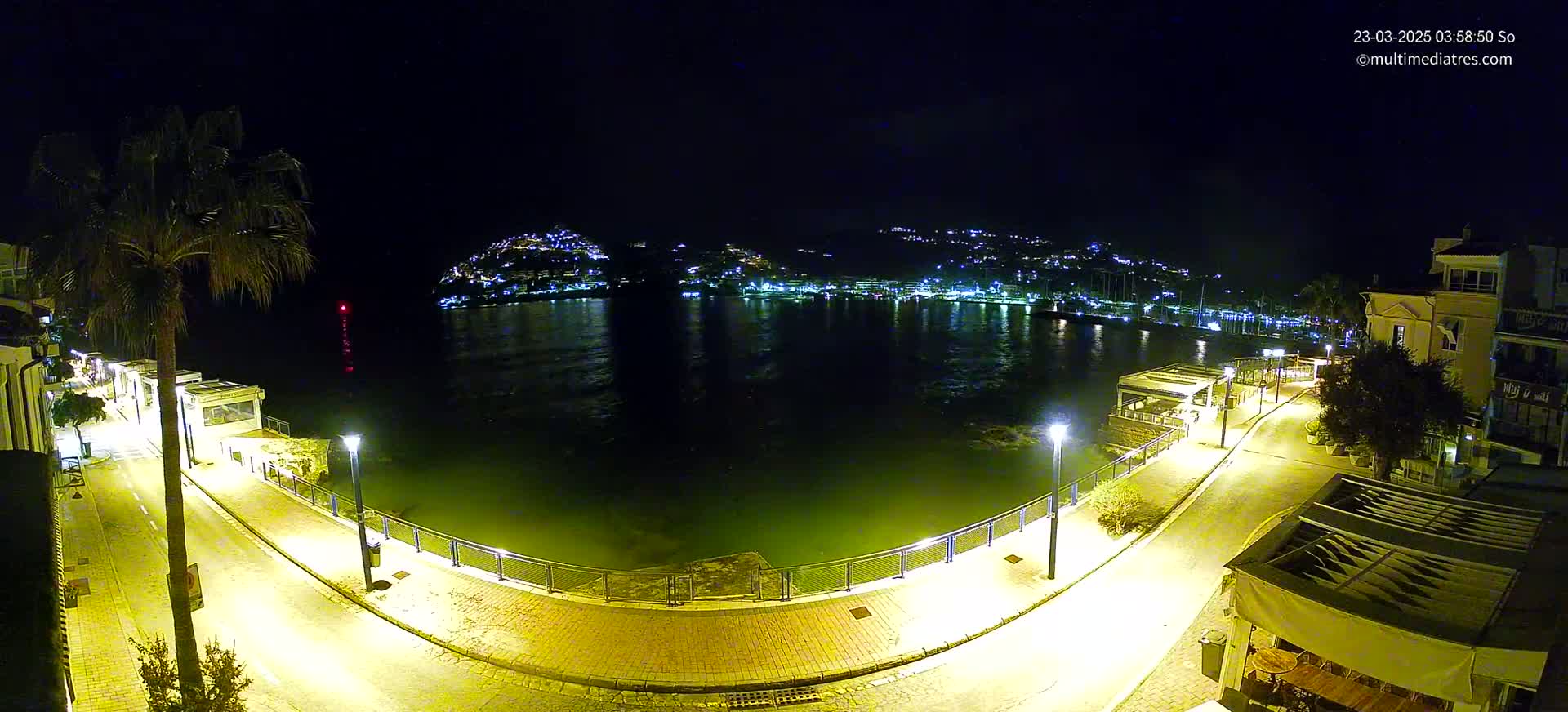 A nighttime aerial view shows a calm body of water reflecting the lights of a town nestled on a hillside, with a paved walkway and palm trees lining the waterfront.