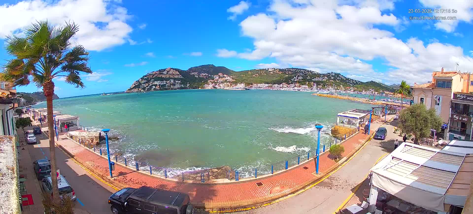 A seaside town with a calm but choppy sea under a partly cloudy, sunny sky is shown from a high vantage point.
