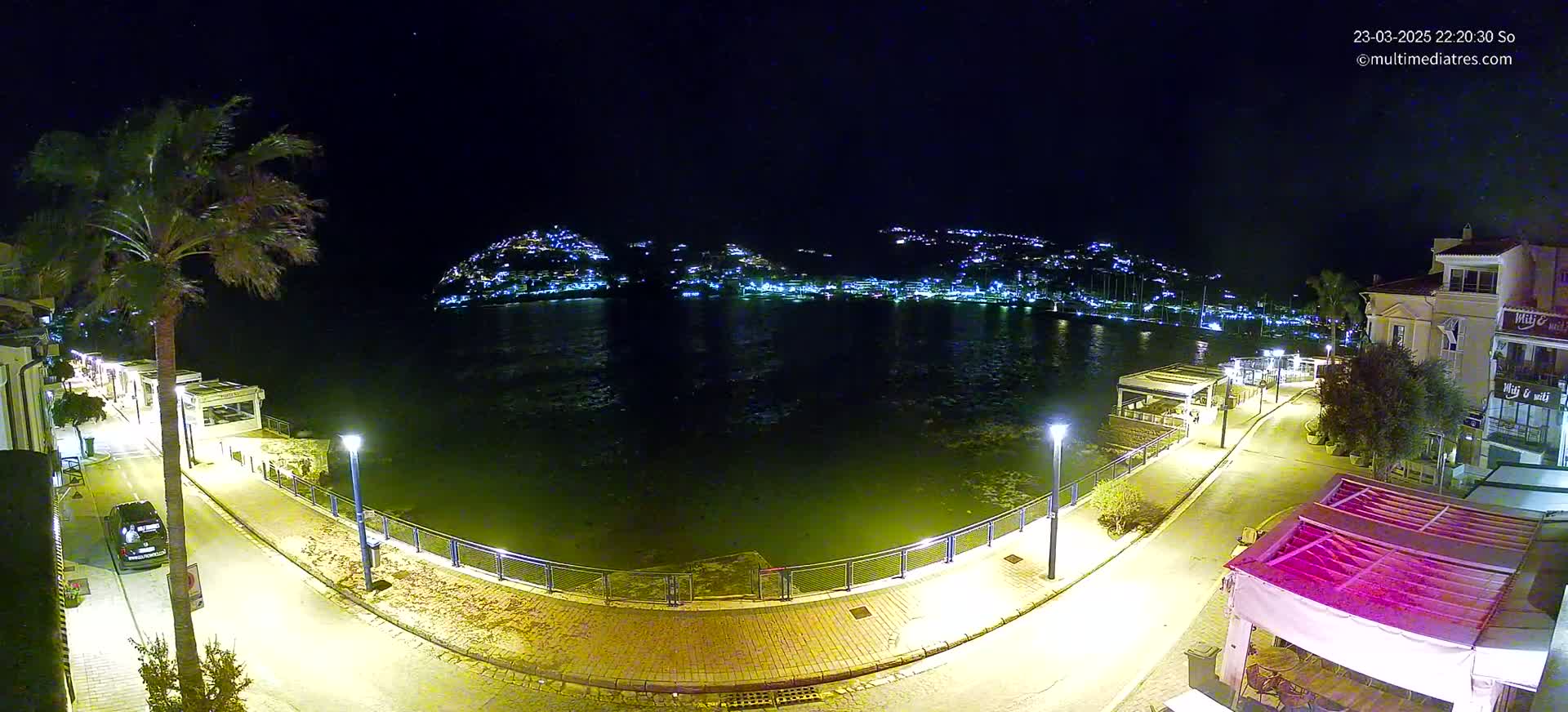 A nighttime, seaside view shows a calm body of water with a town’s lights illuminating the opposite shore, a paved walkway lined with buildings and palm trees, all under a clear, starry sky.