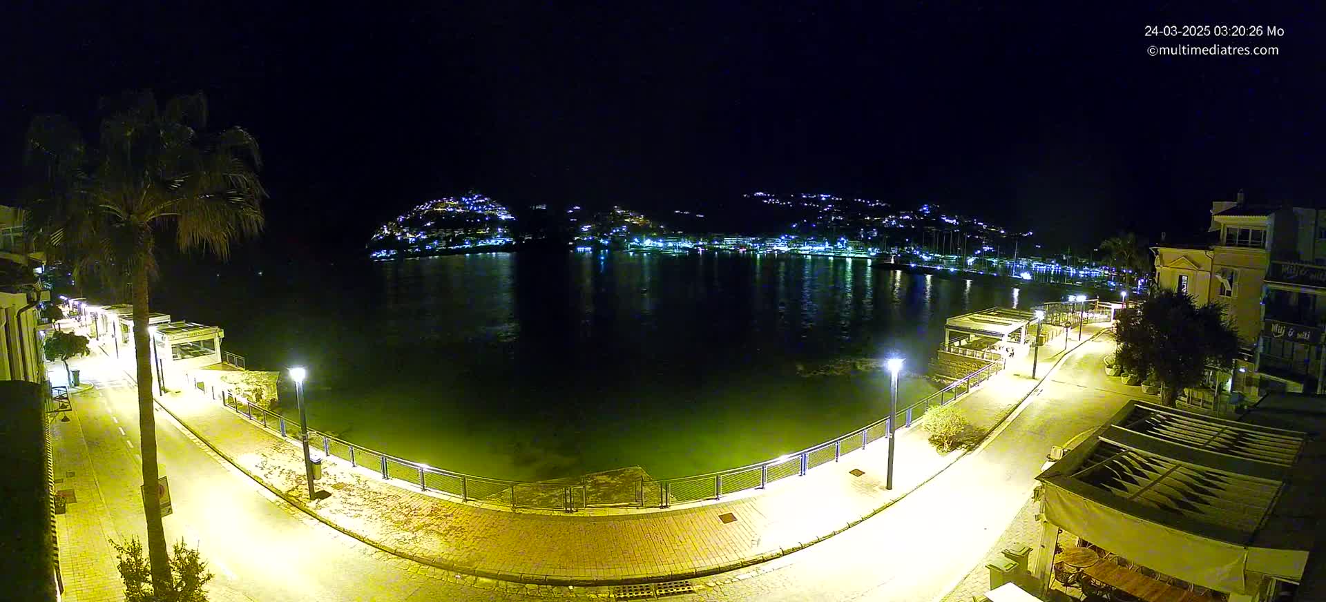 A nighttime, illuminated waterfront scene shows a calm body of water reflecting the lights of a town nestled on a hillside across the water, with a paved walkway and buildings lining the waterfront in the foreground.