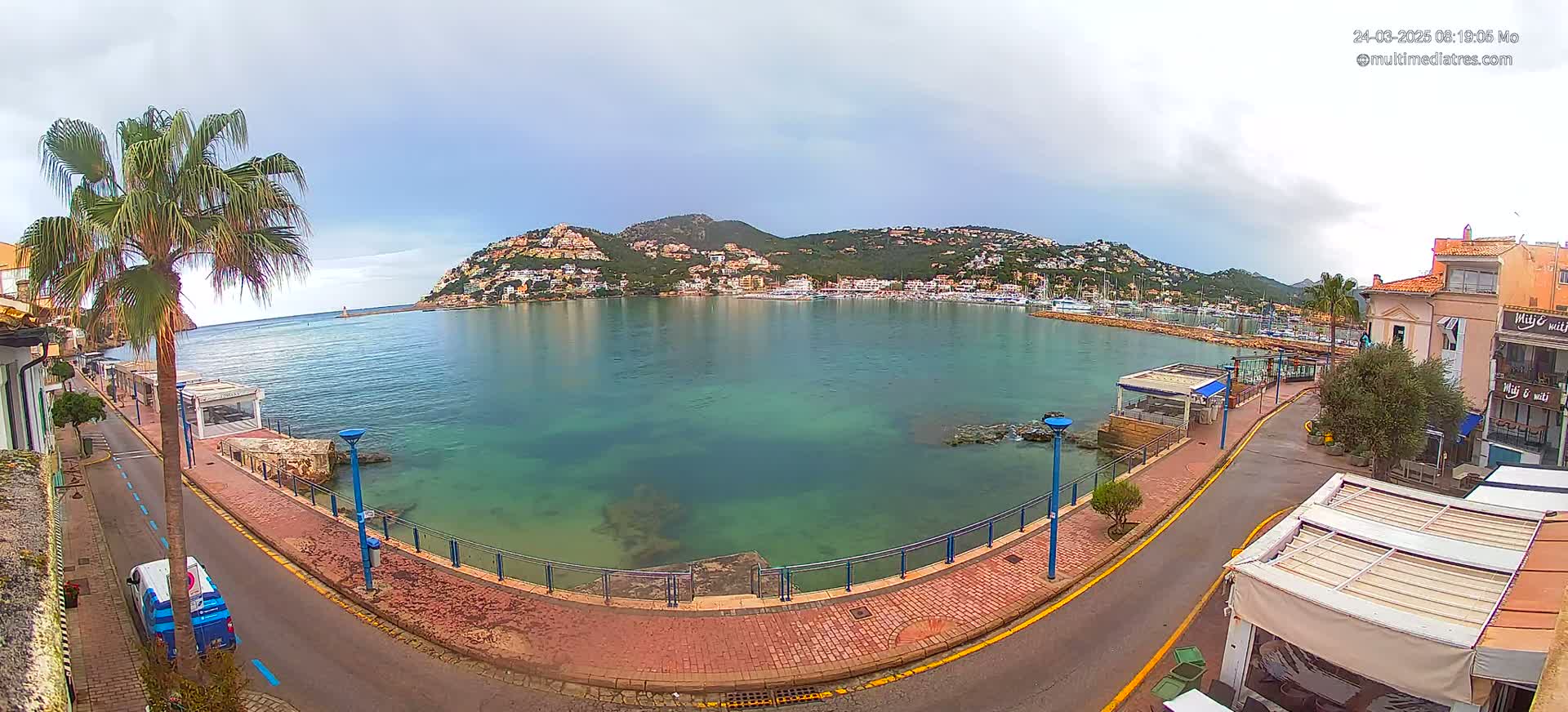 A calm, overcast day overlooks a harbor filled with boats, nestled below a hillside town against a backdrop of mountains.