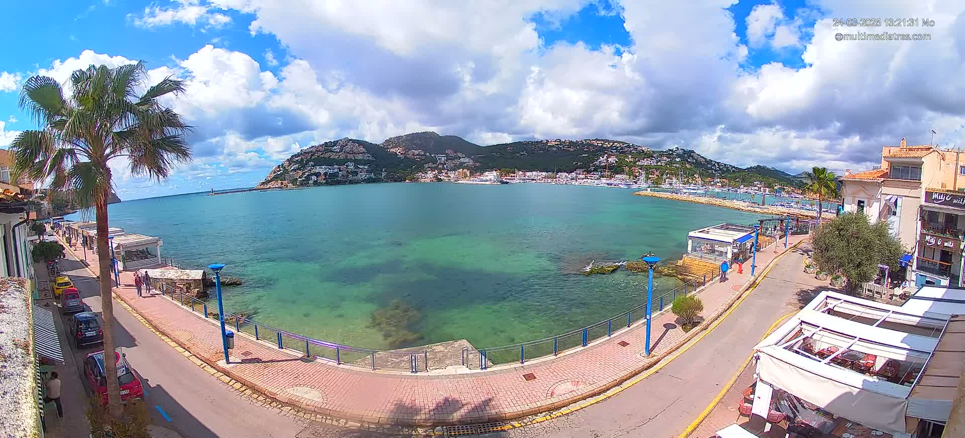 A partly sunny day shows a clear, turquoise bay with a town and a mountain in the background, all viewed from an elevated vantage point along a paved waterfront walkway.