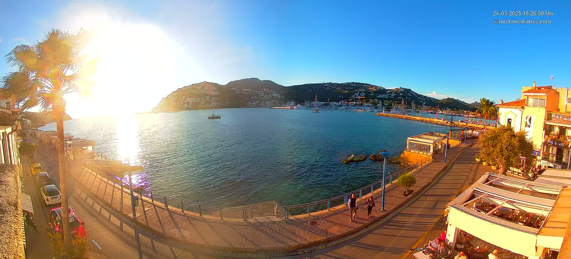 A seaside town at sunset, with a calm harbor filled with boats and buildings lining a paved walkway.