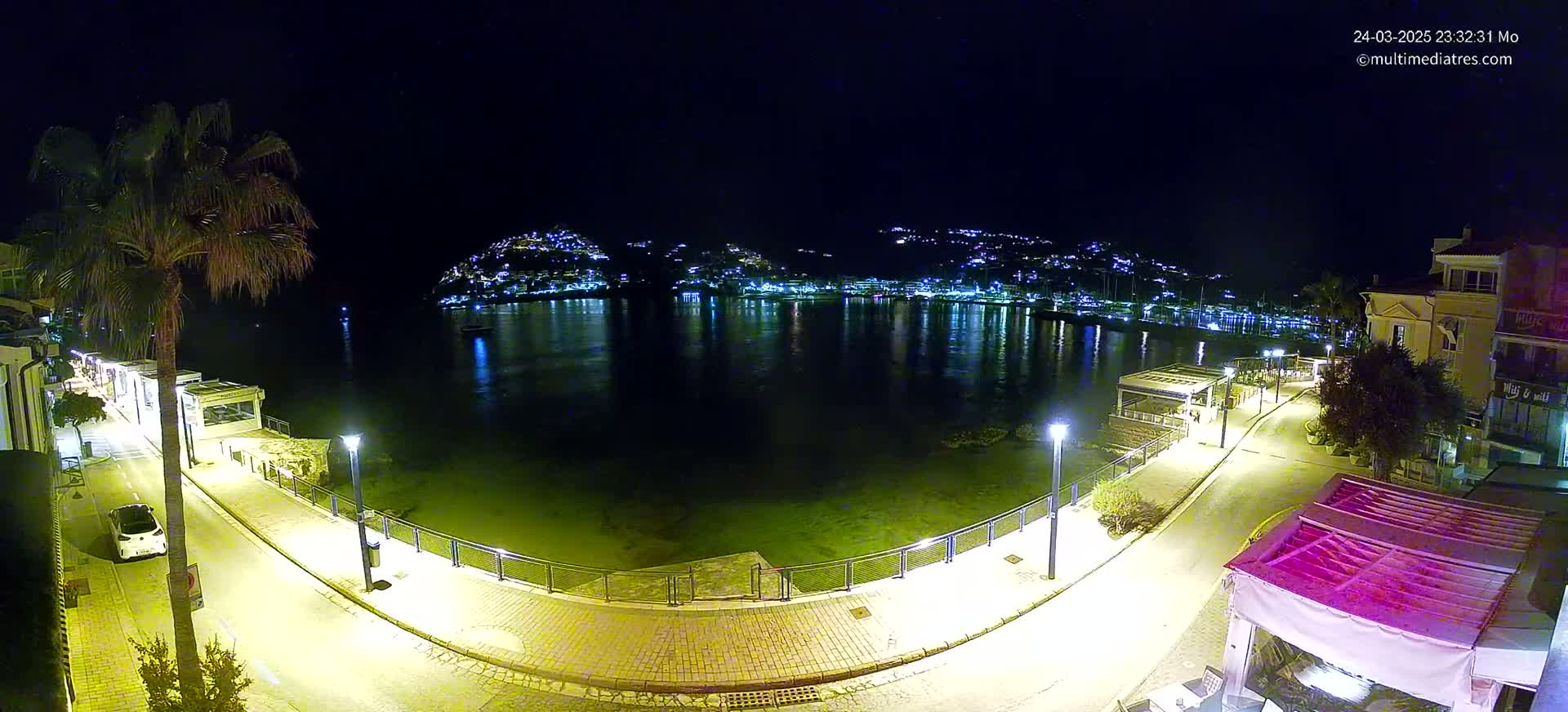 A nighttime view of a calm bay, illuminated by lights lining a paved waterfront path, with a hillside town glowing in the distance.