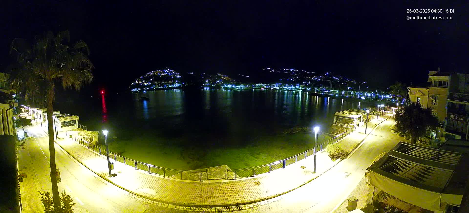 A nighttime, illuminated waterfront scene shows a calm bay reflecting the lights of a town nestled on a hillside, with a paved walkway and buildings lining the shore.