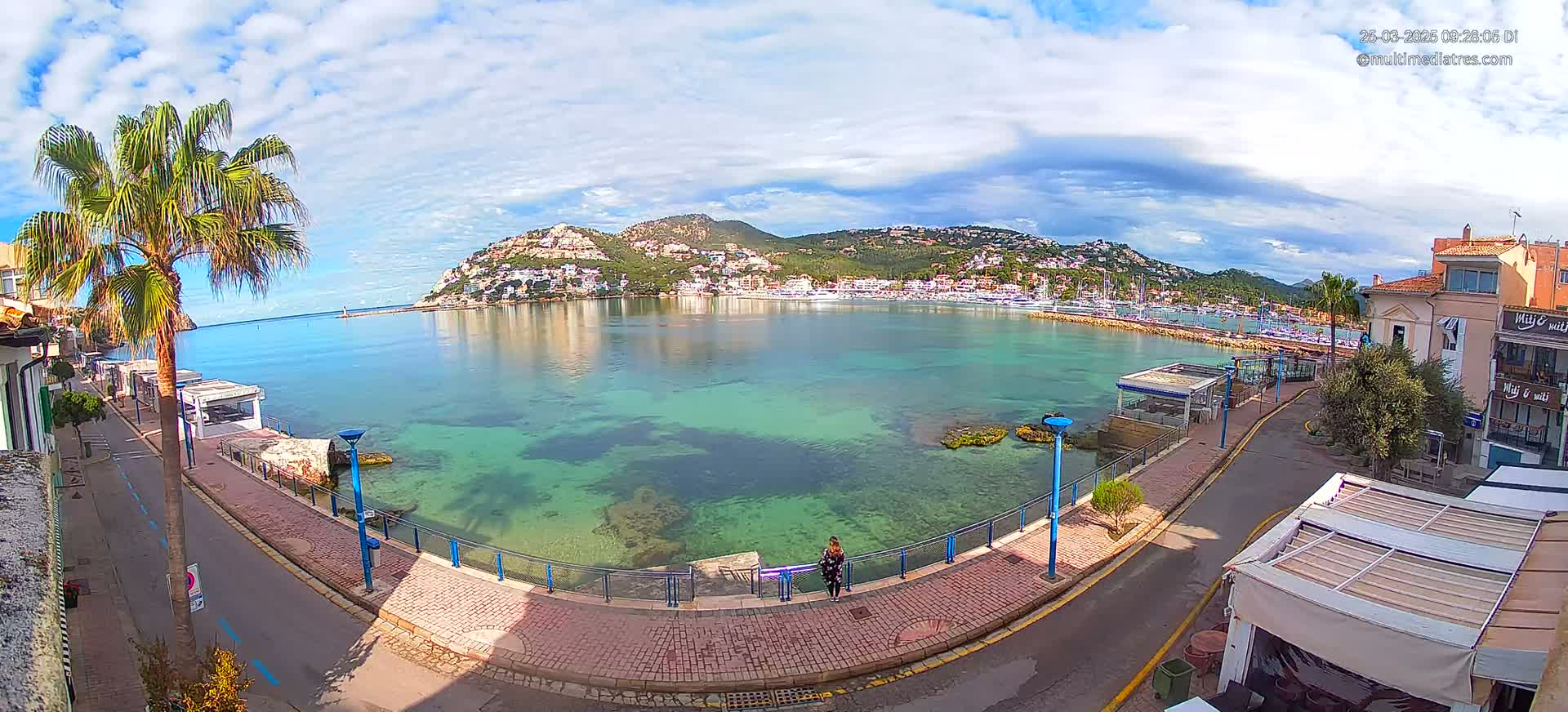 A partly cloudy day reveals a calm, clear turquoise bay with a town and hills in the background, viewed from a seaside promenade.