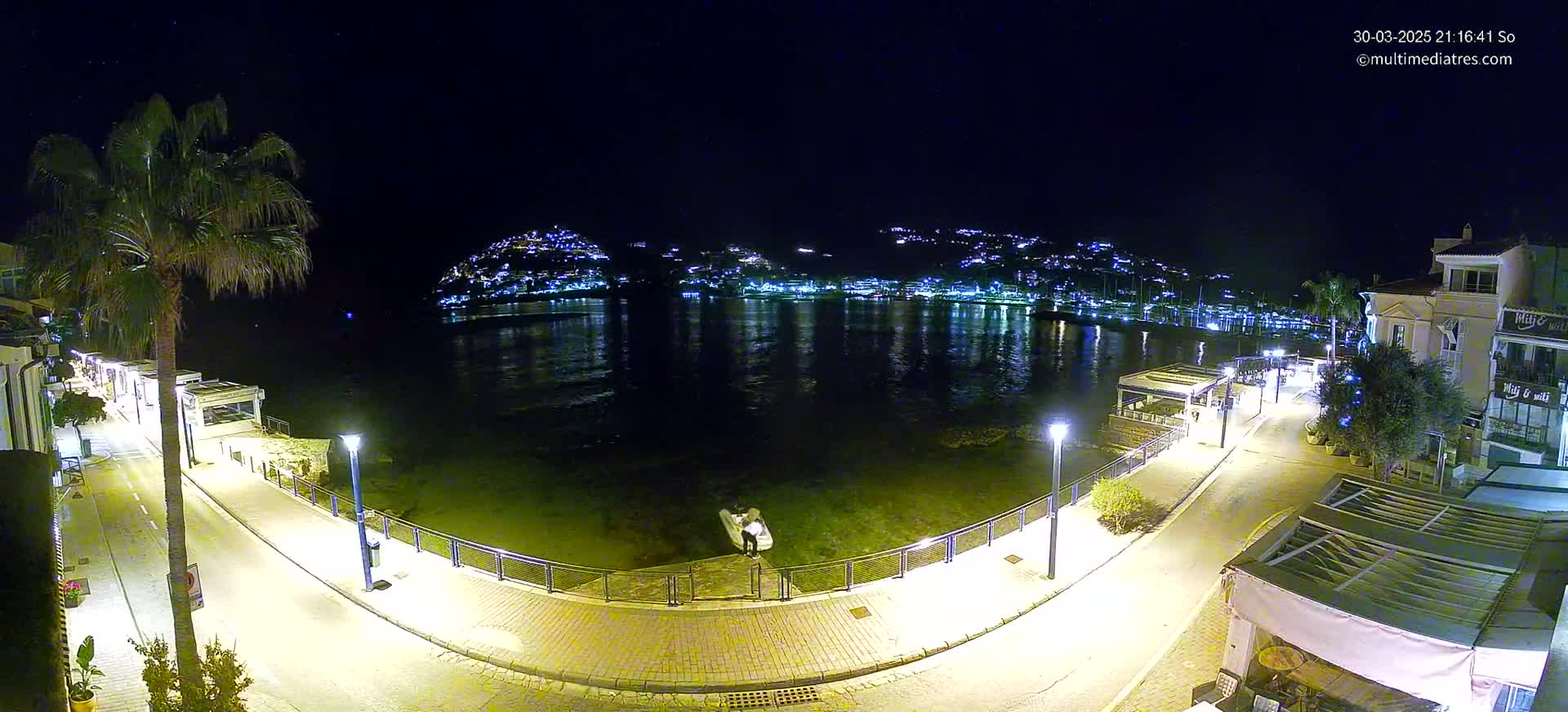 A nighttime view of a calm body of water reflecting the lights of a town nestled on a hillside, with a paved walkway and palm trees lining the waterfront.