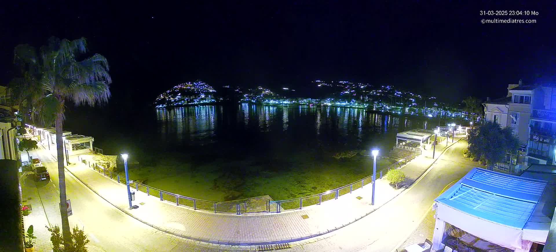 A nighttime view of a waterfront promenade, with a calm body of water reflecting the lights of buildings on a hill in the background, under a clear sky.
