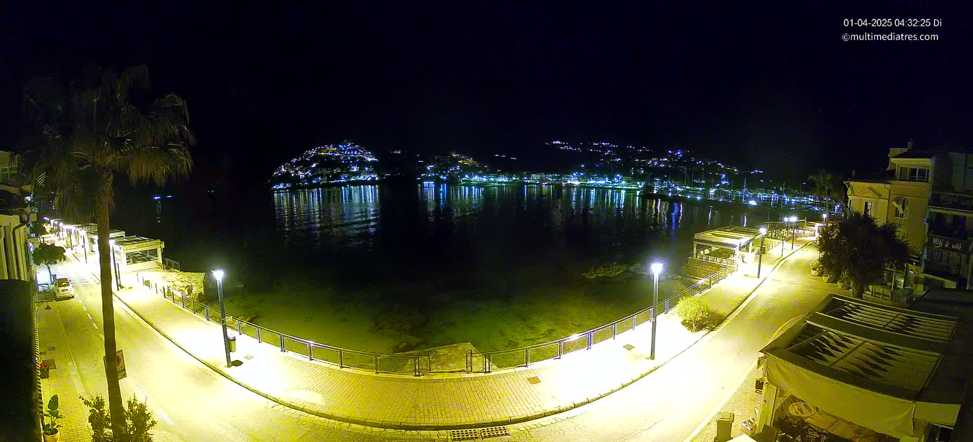 A nighttime, waterfront scene shows a paved walkway illuminated by streetlights curving around a calm body of water reflecting the lights of a town on the opposite shore.