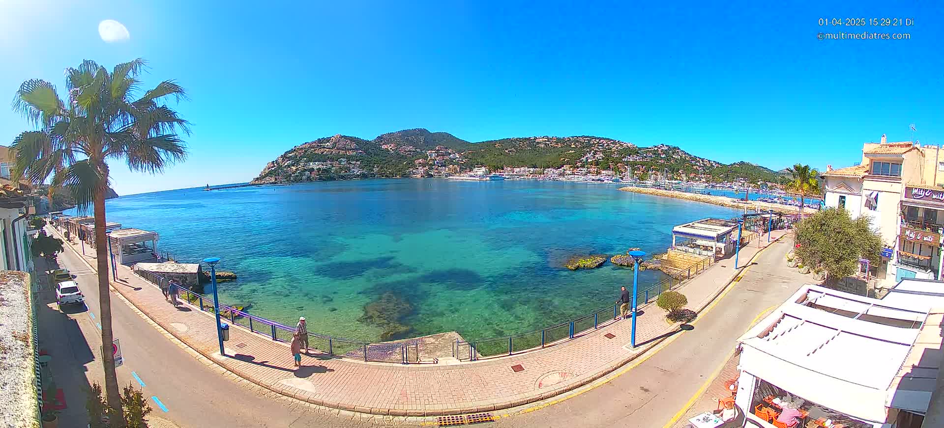 A sunny day overlooking a clear, turquoise bay with a town and hillside buildings in the background, viewed from a seaside promenade.