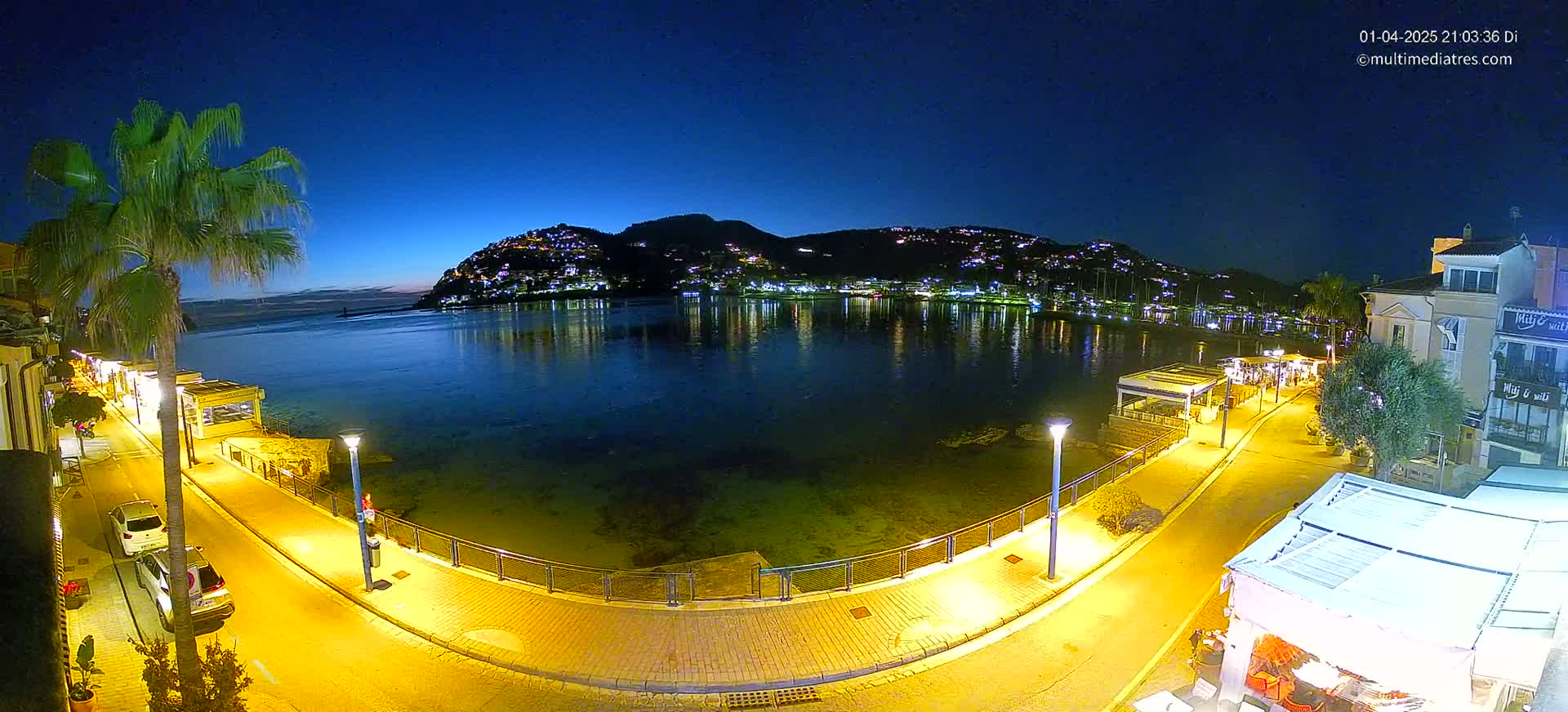 A nighttime view of a calm bay, illuminated by streetlights, with a hilltop town reflecting in the water and a paved waterfront path lined with buildings and palm trees.