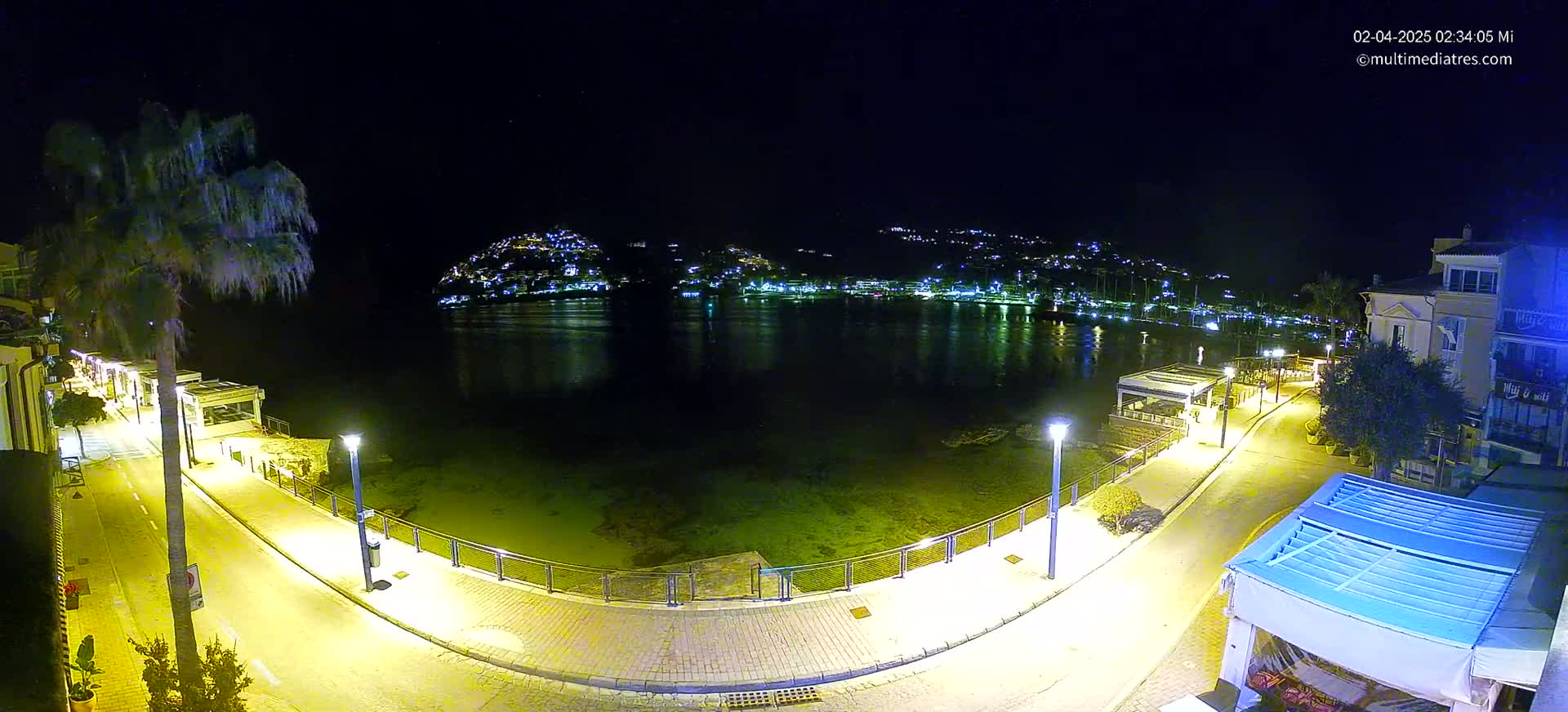 A nighttime, illuminated waterfront scene shows a calm body of water reflecting a hilltop town, with a paved walkway and buildings lining the shore.