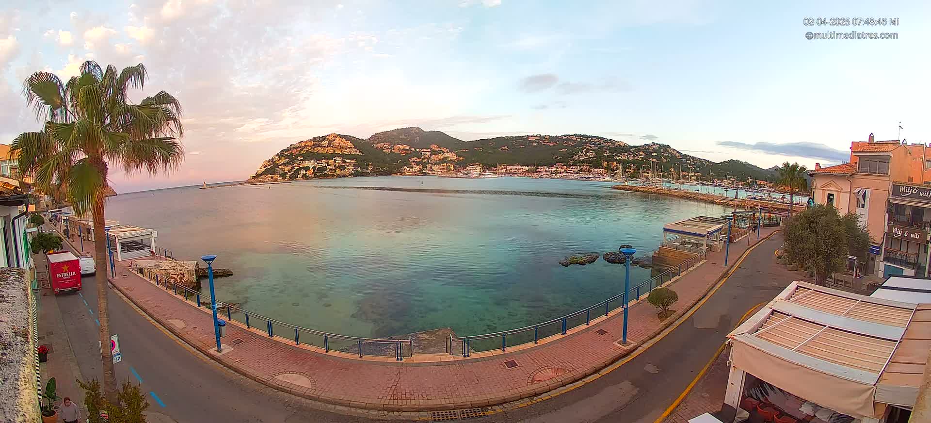 A calm bay with clear, shallow water near a waterfront promenade lined with buildings and palm trees under a partly cloudy sky.