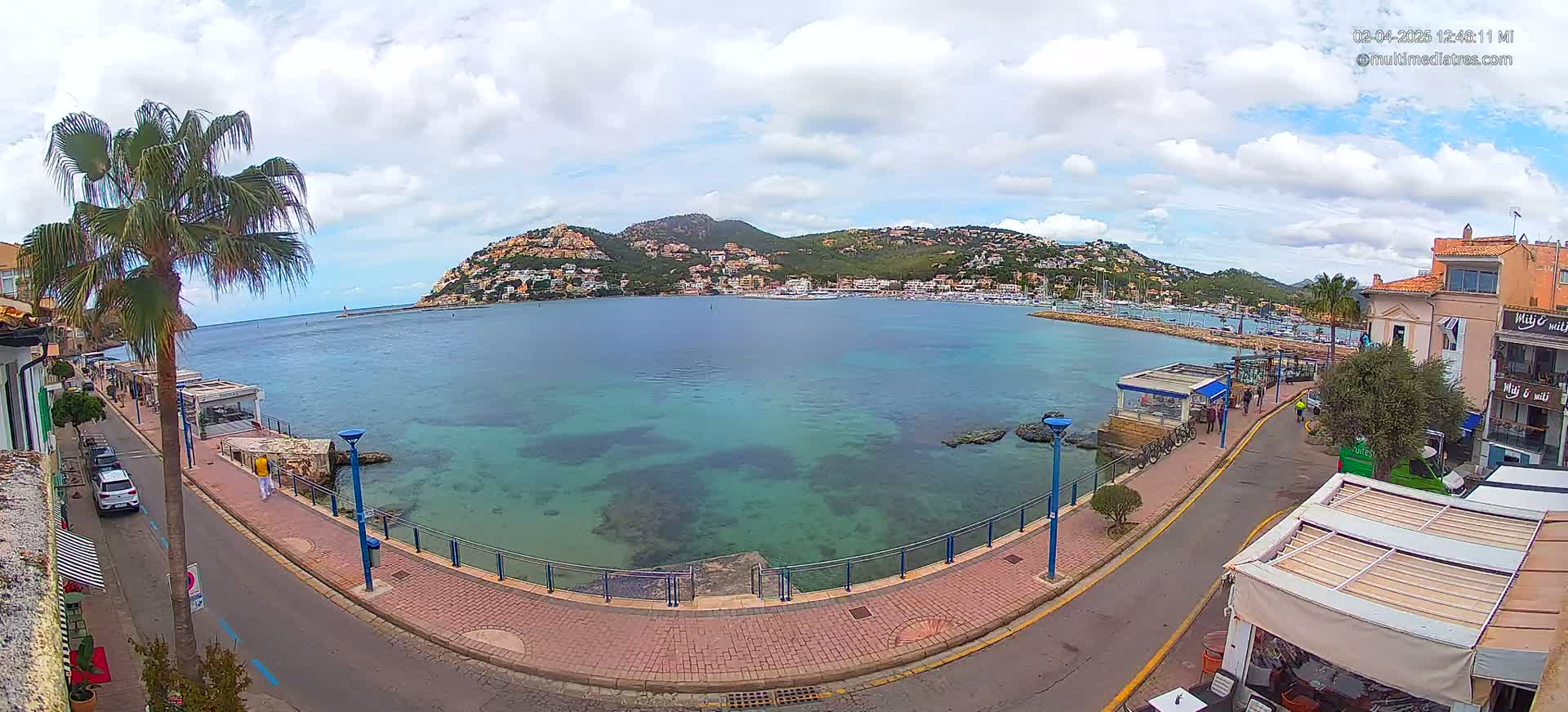 A partly cloudy day overlooking a calm, clear turquoise bay with a town and hillside buildings in the background, seen from a waterfront road with palm trees and buildings lining the shore.