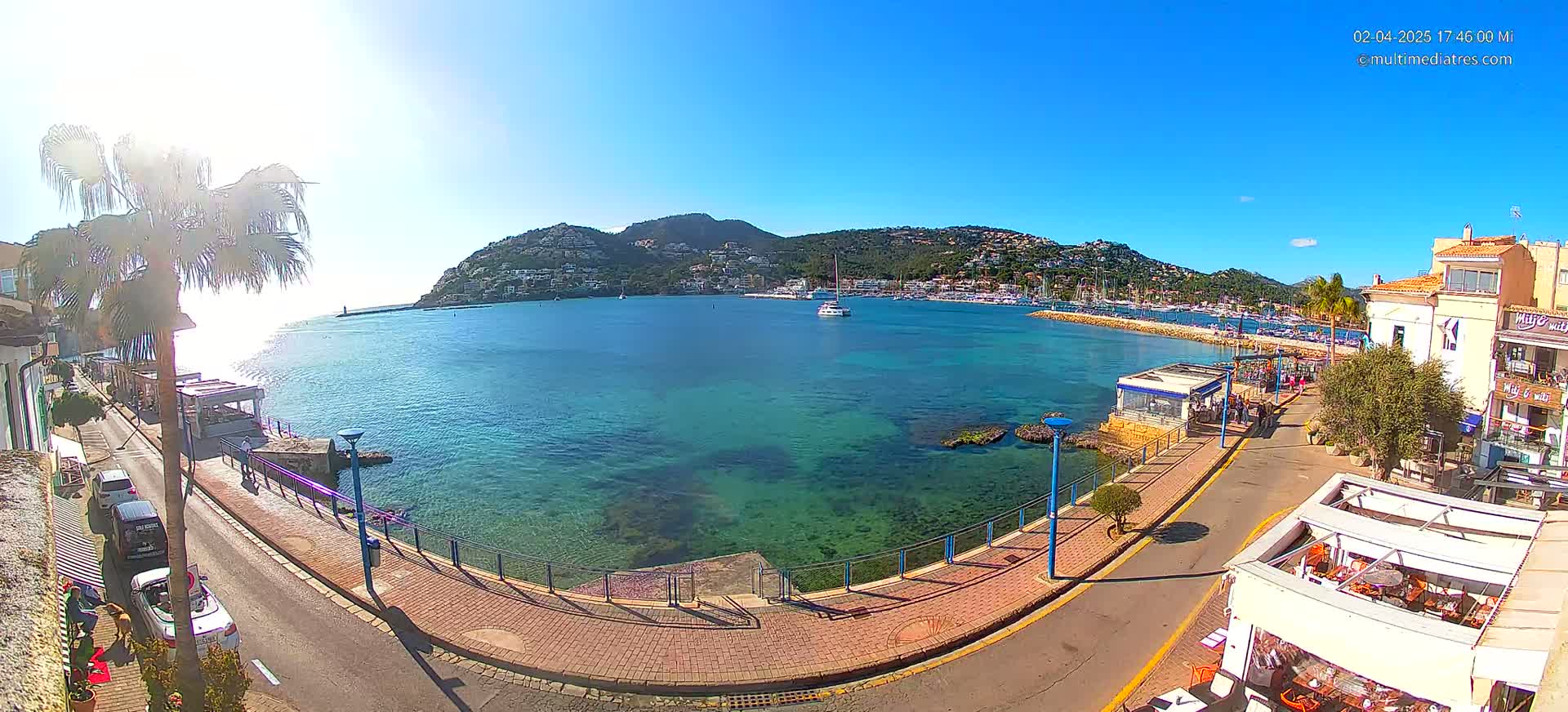A sunny day overlooking a calm, clear turquoise bay with boats in a marina, a paved walkway, and buildings along the shore.