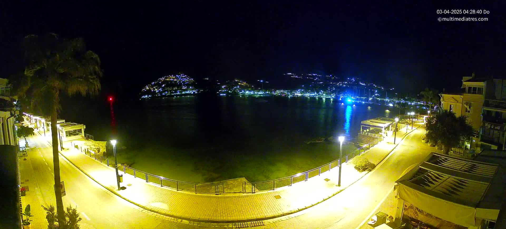 A nighttime view of a calm harbor, with illuminated walkways and buildings lining the shore, under a clear, dark sky.