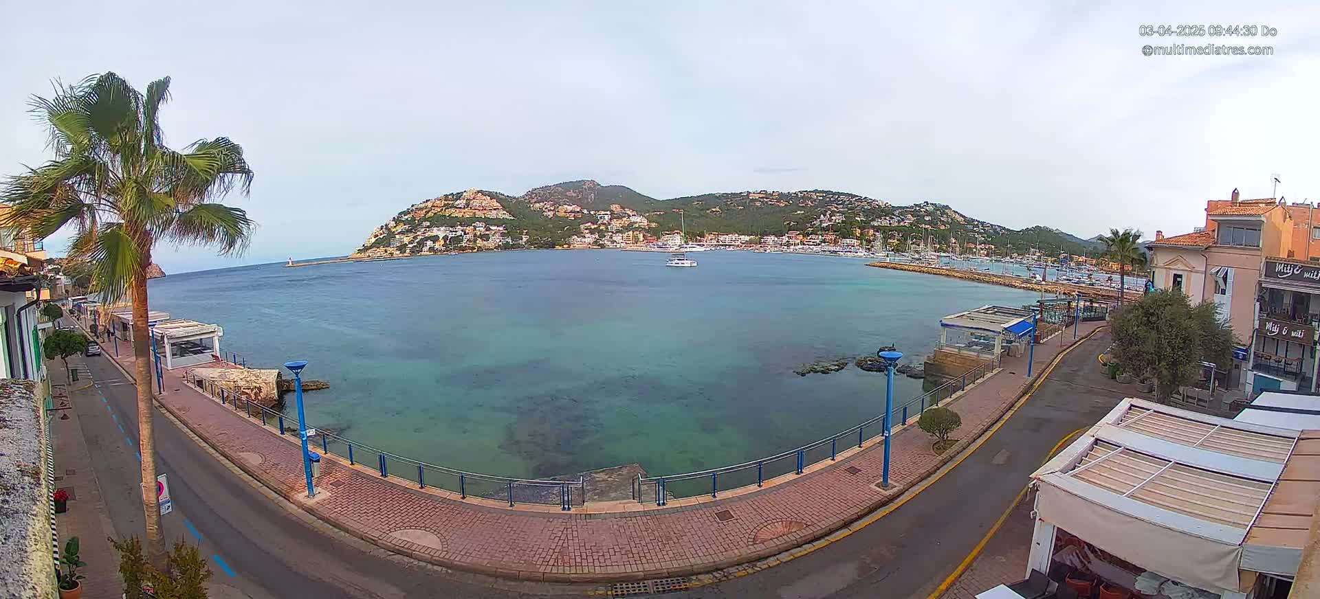 A calm, overcast day reveals a coastal town's harbor, showcasing a small marina with several boats and a hill dotted with buildings in the background.