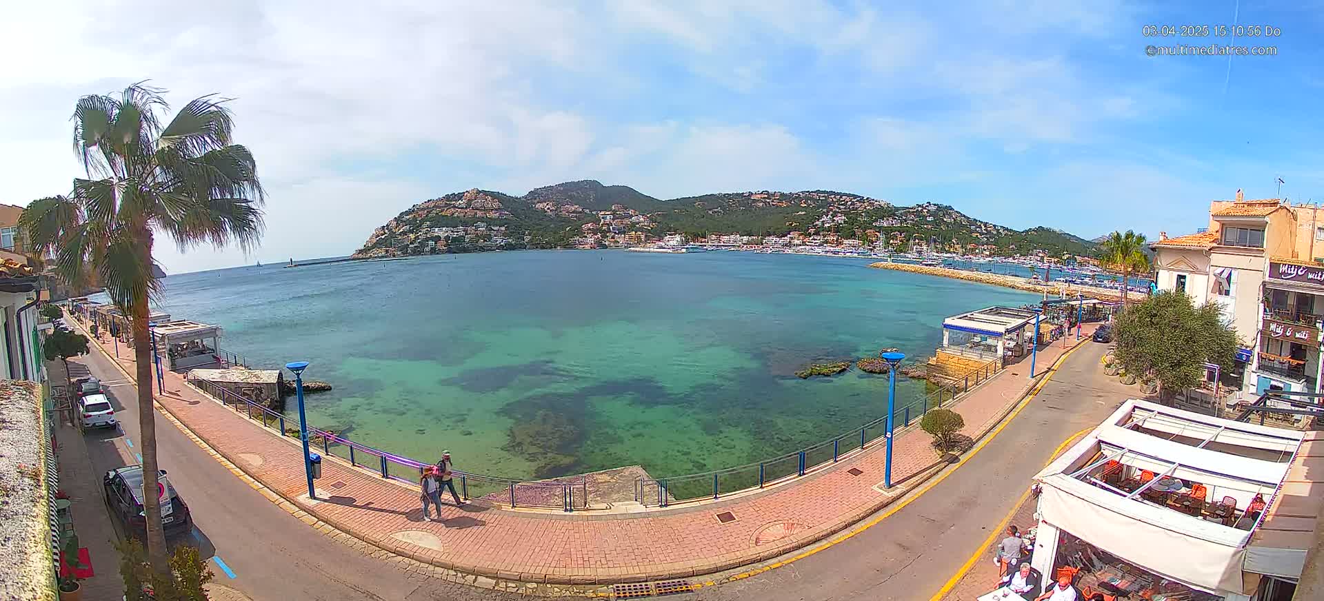 A seaside town with clear, shallow turquoise water, a paved walkway, parked cars, and buildings, under sunny skies.