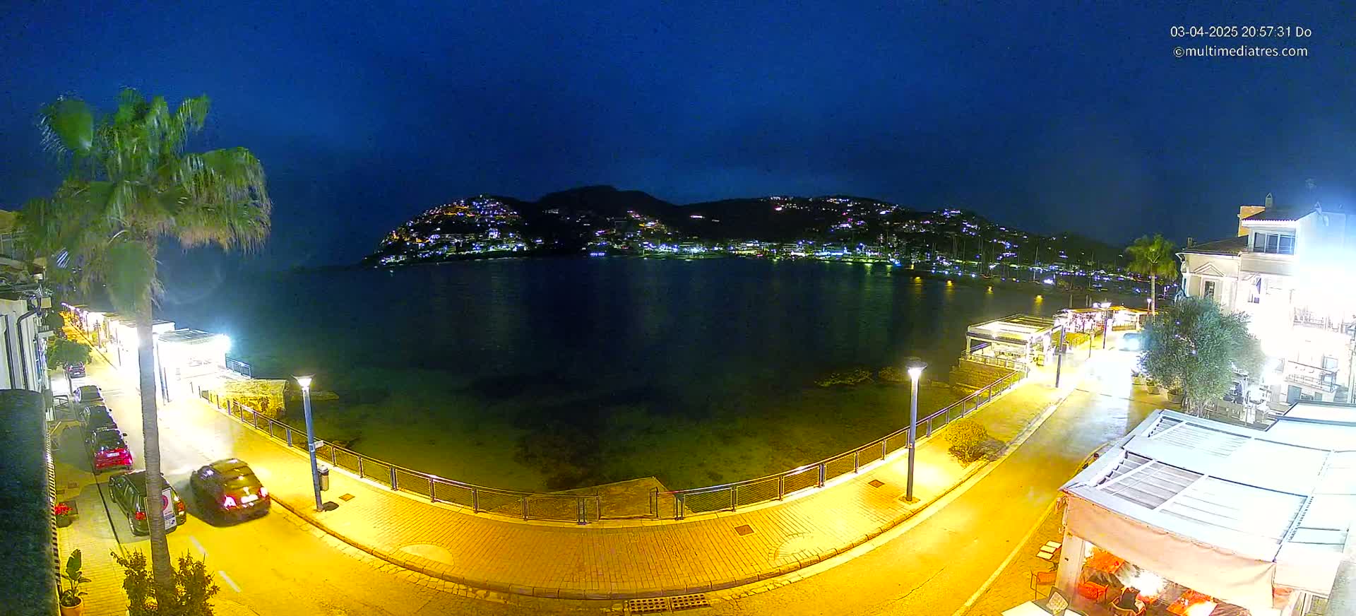 A nighttime view of a calm bay, illuminated by streetlights, with a hill dotted with lights in the background, and a paved waterfront walkway with cars parked along it.